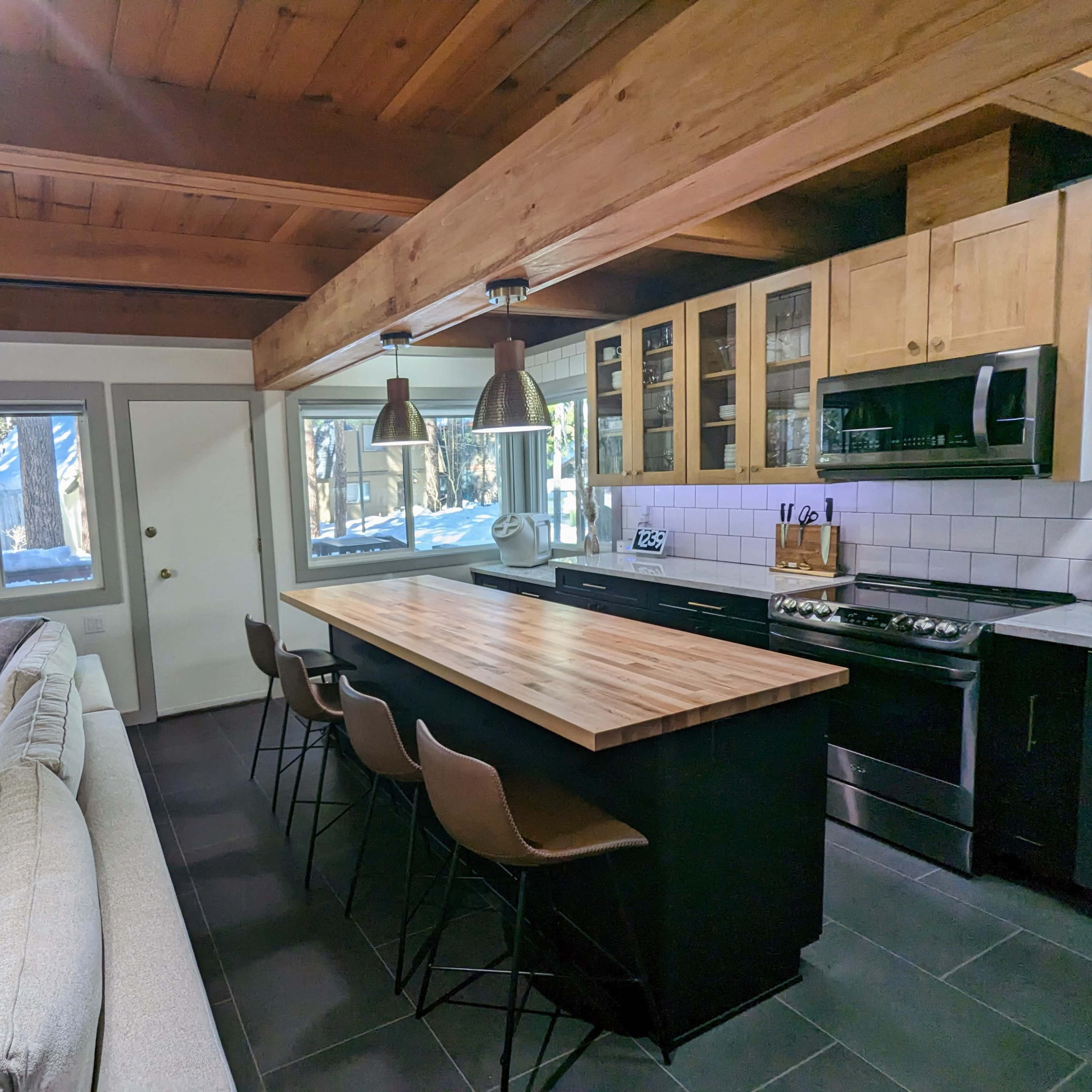 A modern kitchen with a wooden island, bar stools, and stainless steel appliances, situated beside a cozy living area featuring a beige sofa.