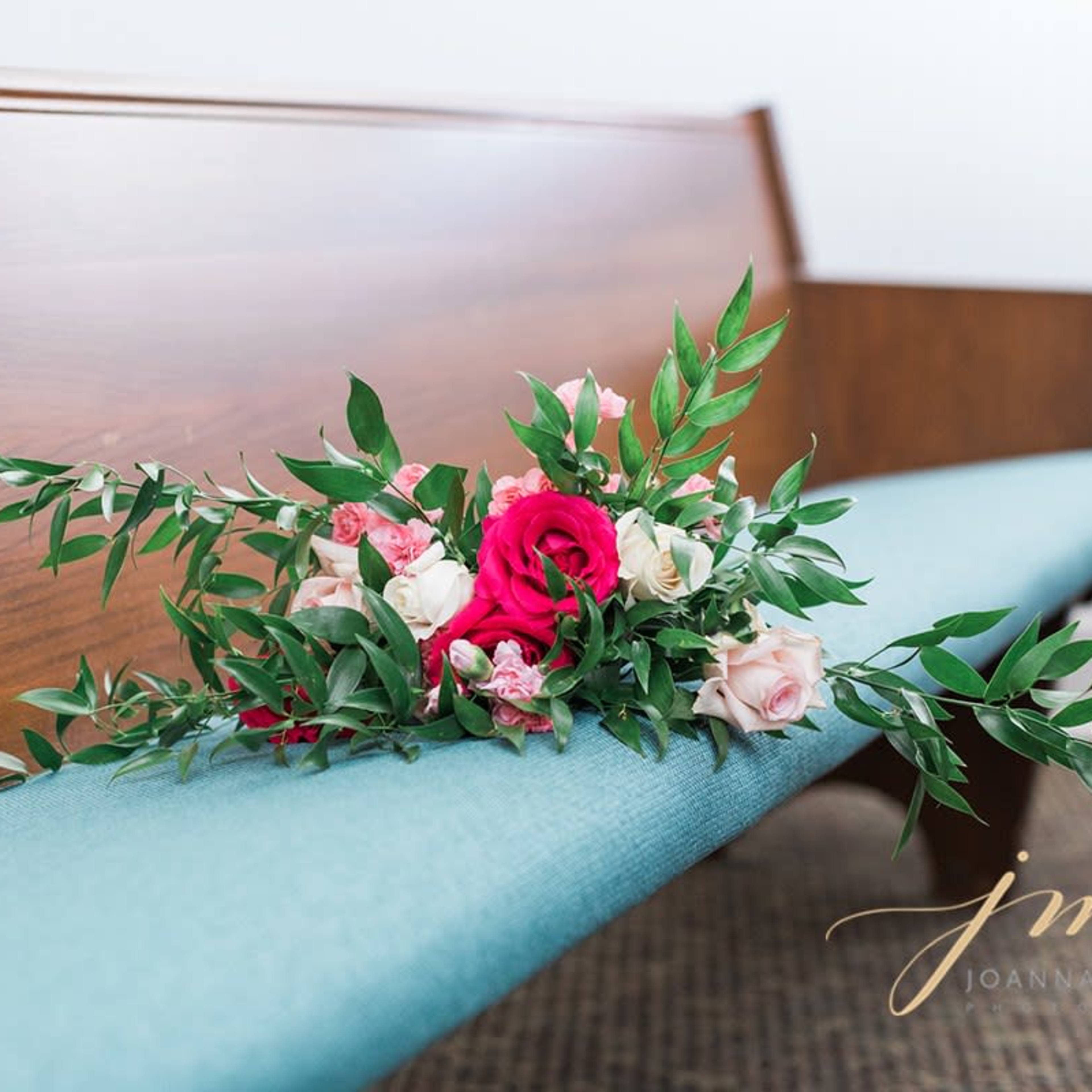 A floral arrangement with pink and white roses is placed on a wooden pew with a teal cushion.