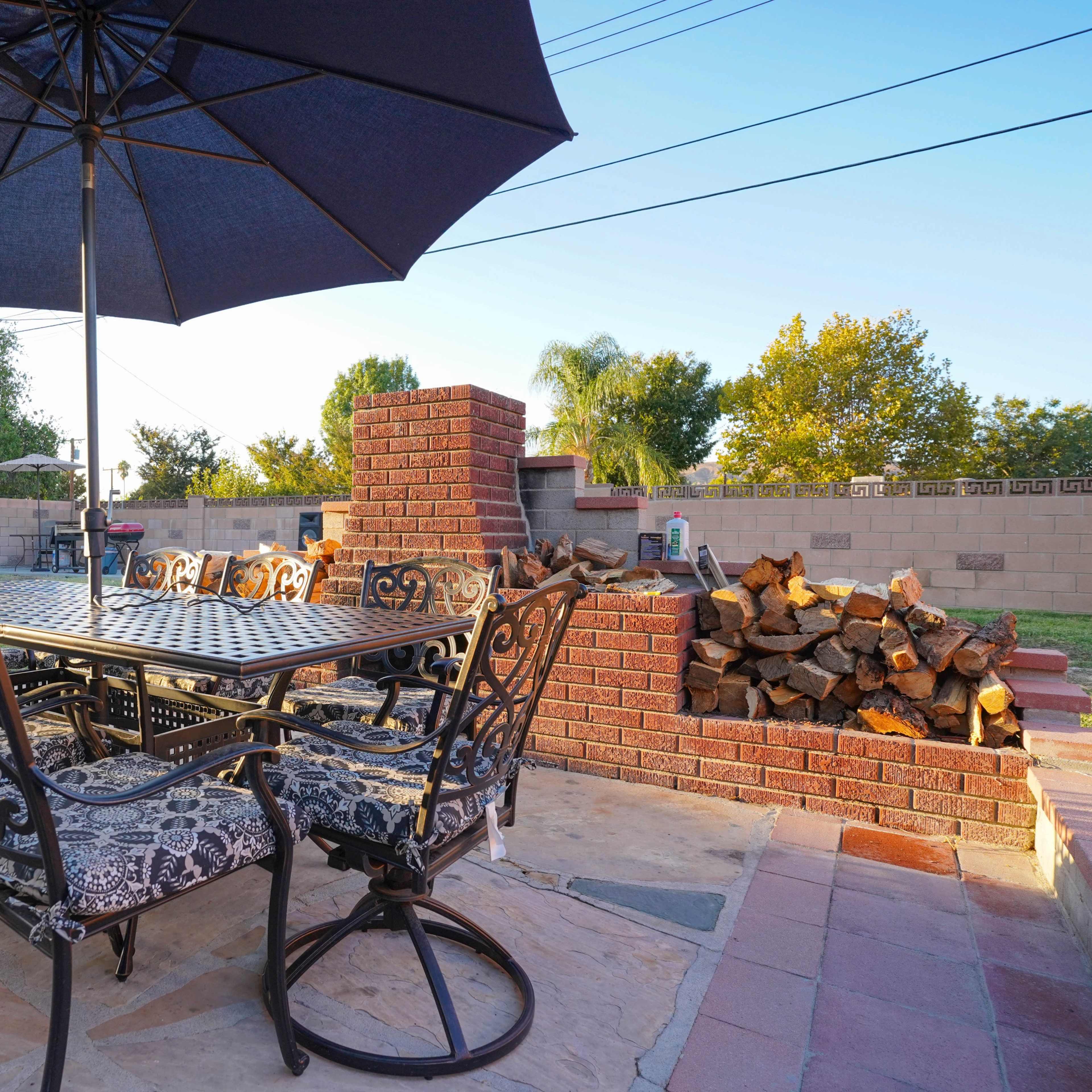 A covered outdoor dining table with chairs is set beside a brick fireplace and a stacked pile of firewood in a landscaped backyard.