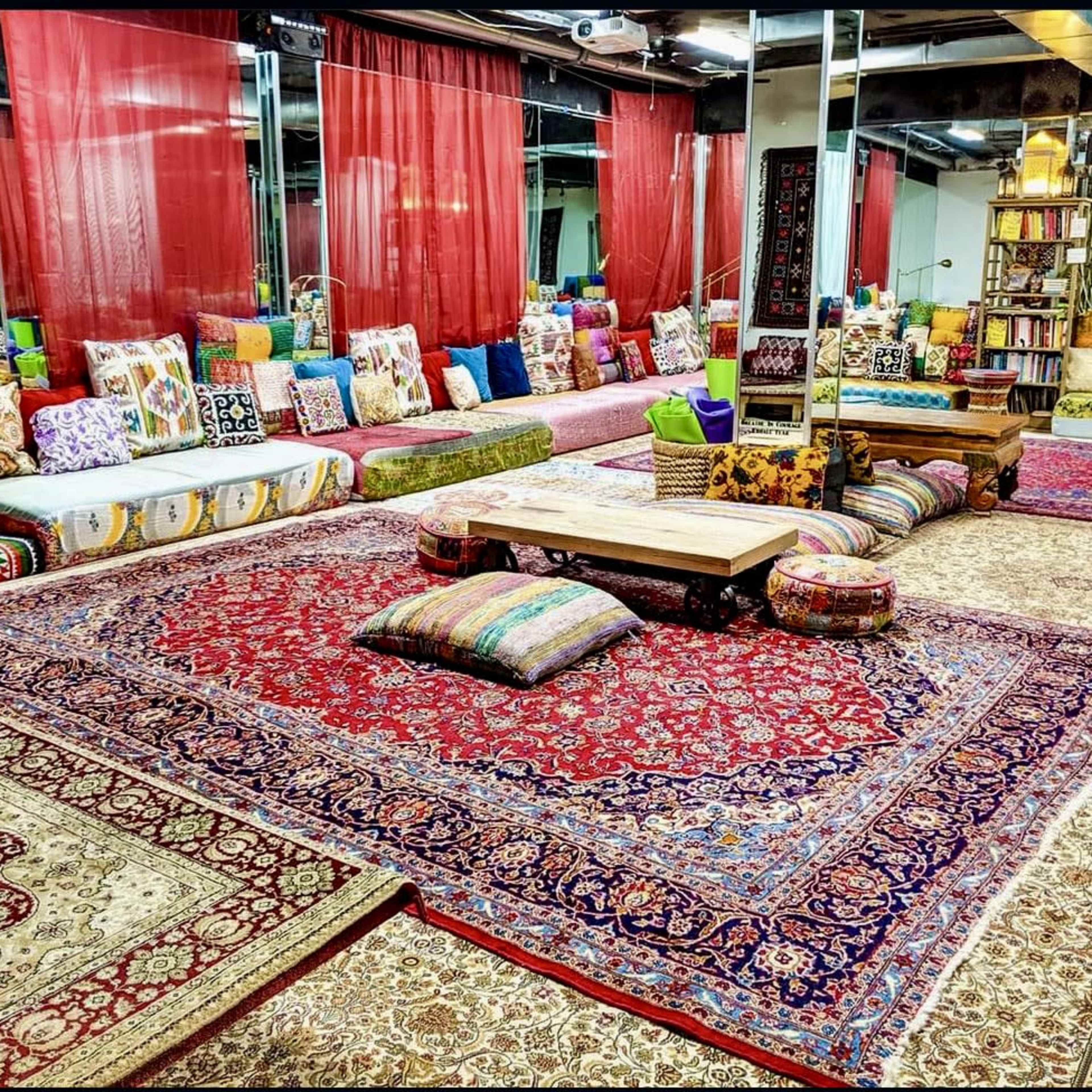The room features an array of colorful cushions and patterned rugs arranged around a central wooden table, with sheer red drapes and mirrored walls enhancing the inviting atmosphere.
