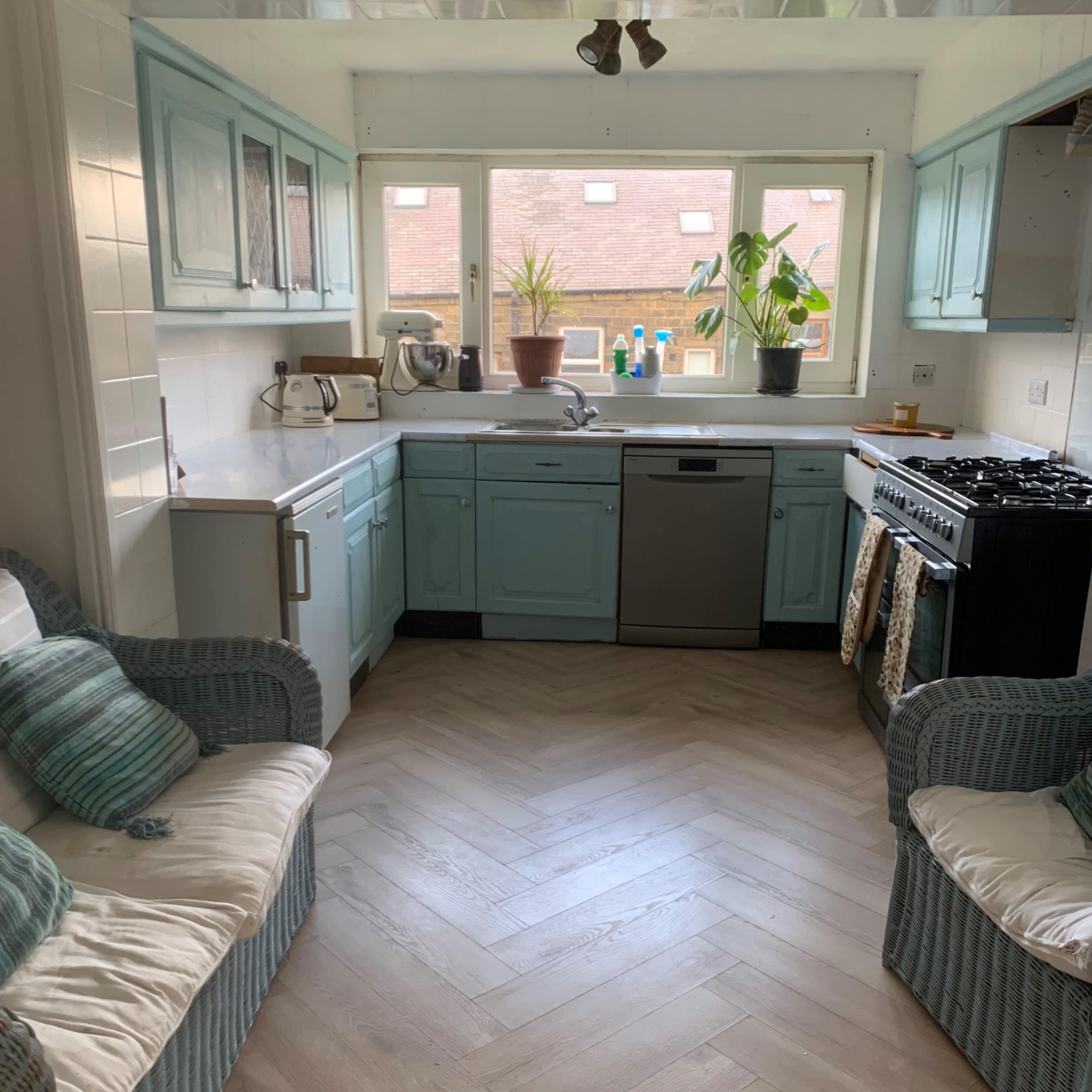 A kitchen featuring turquoise cabinets, a stainless steel dishwasher, a gas stove, and two wicker chairs with cushions positioned near a window.