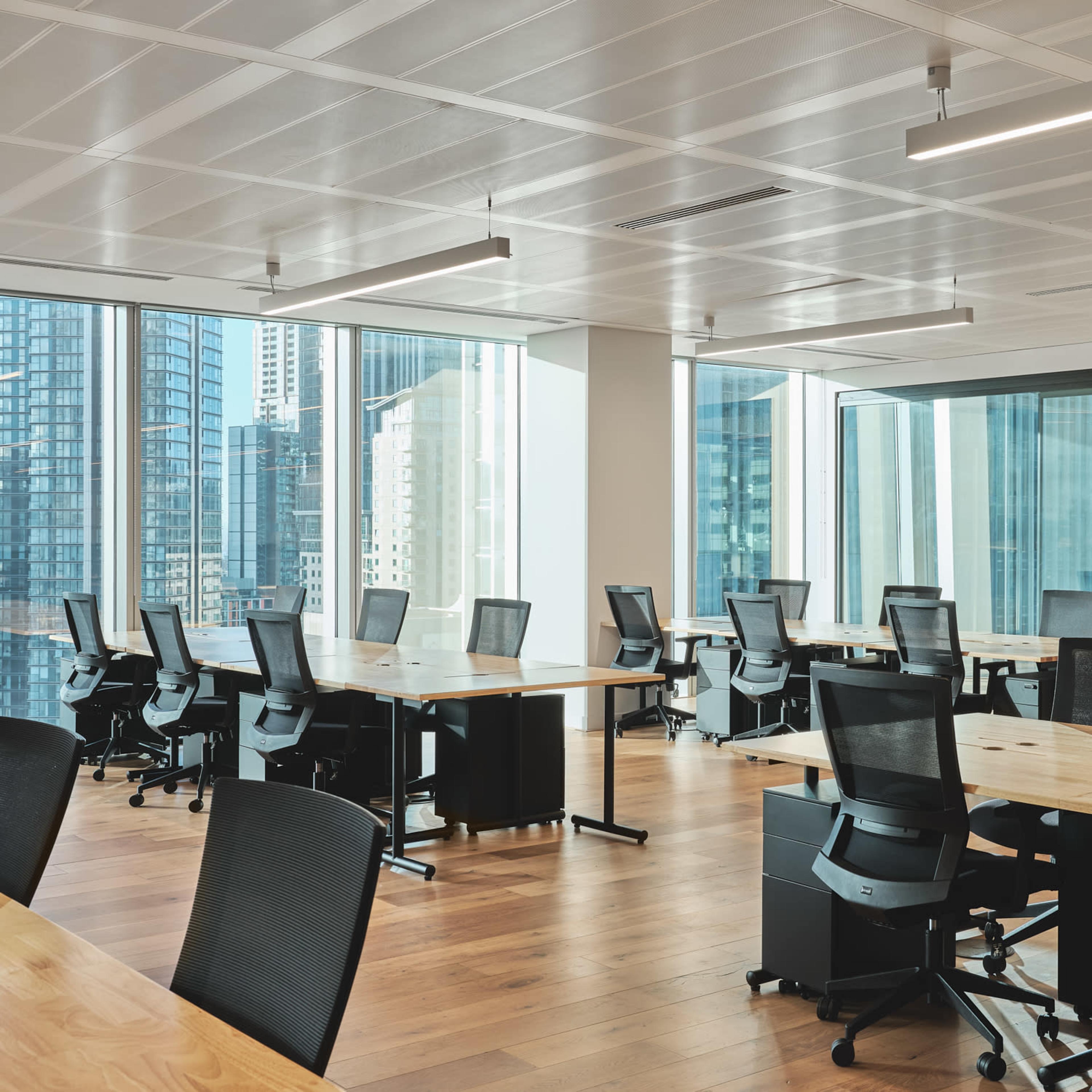 A modern office space with several workstations featuring black chairs and wooden desks, surrounded by large windows that provide a view of a city skyline.