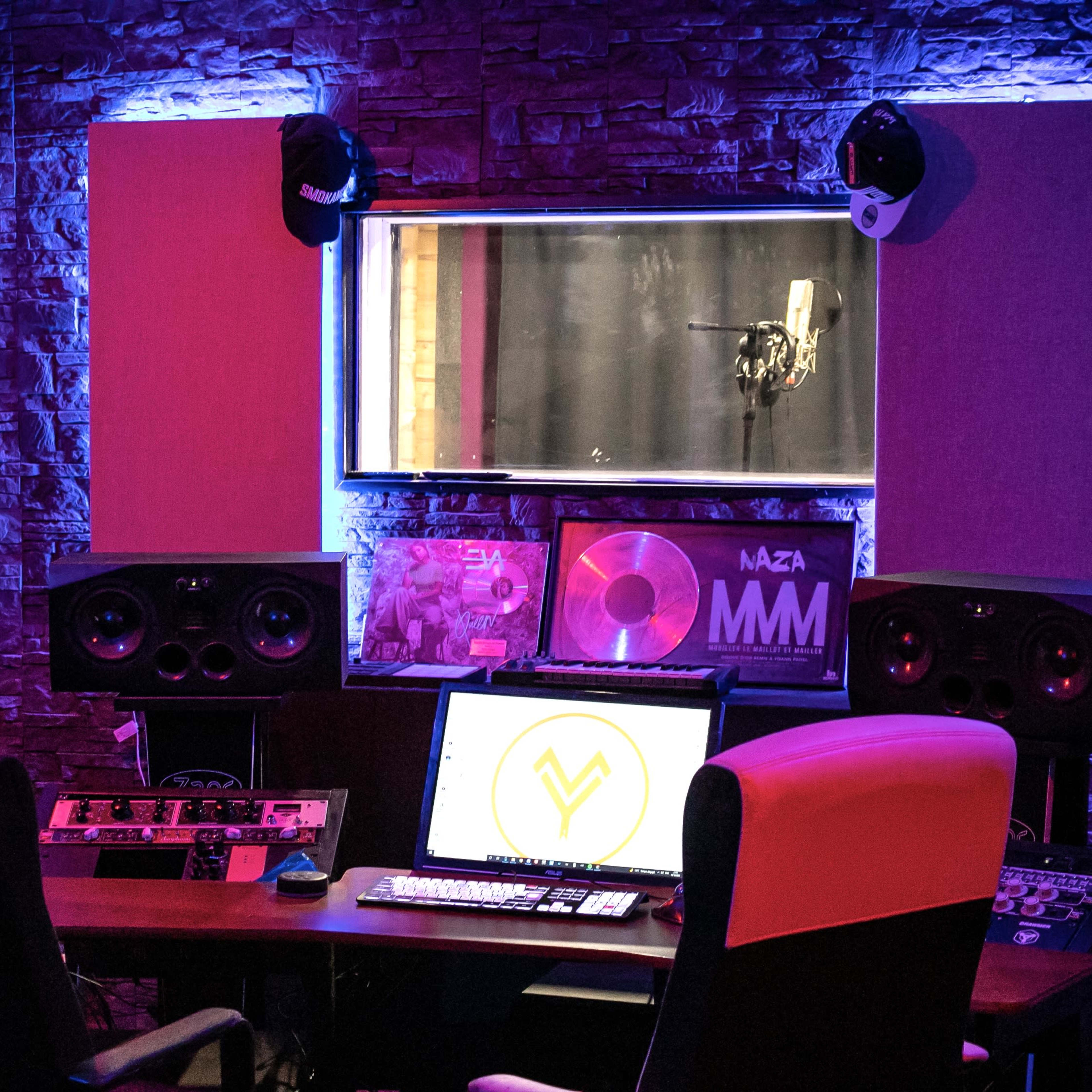 A modern music studio with a microphone in a soundproof booth, large speakers, and a computer setup on a wooden desk.