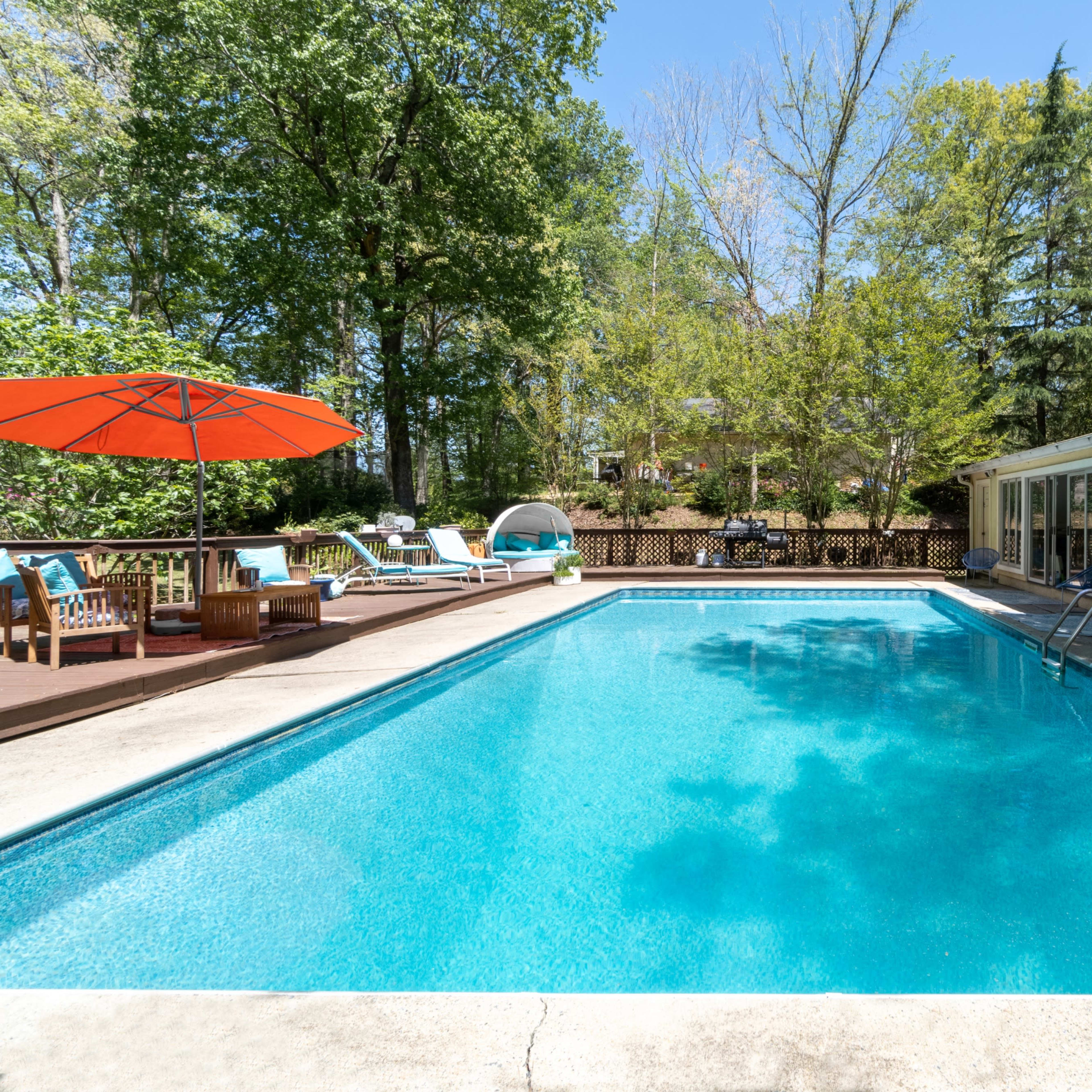 The scene features a clear swimming pool surrounded by lounge chairs, a red umbrella, and a house in a wooded backyard.