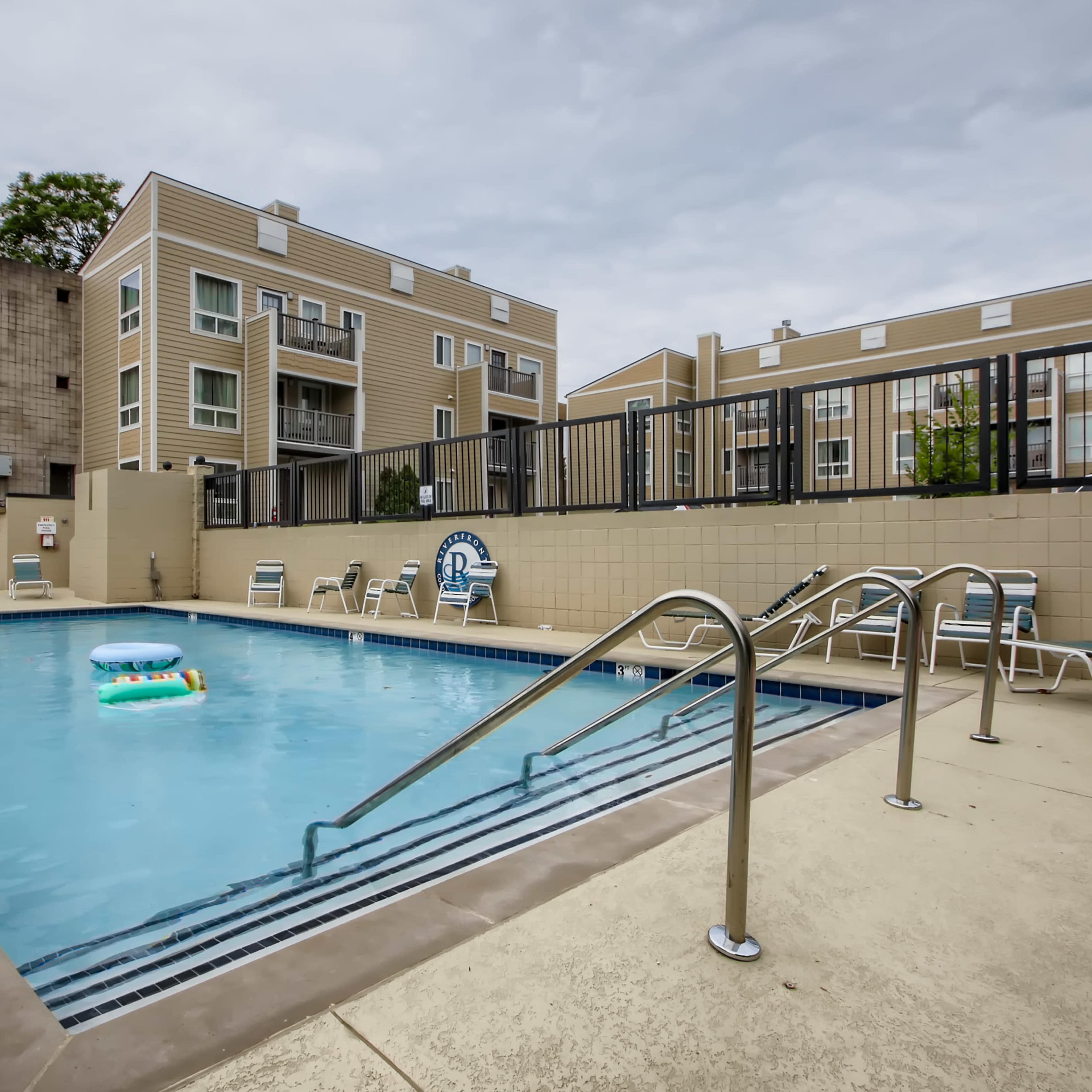 A swimming pool surrounded by lounge chairs and apartment buildings against a cloudy sky.