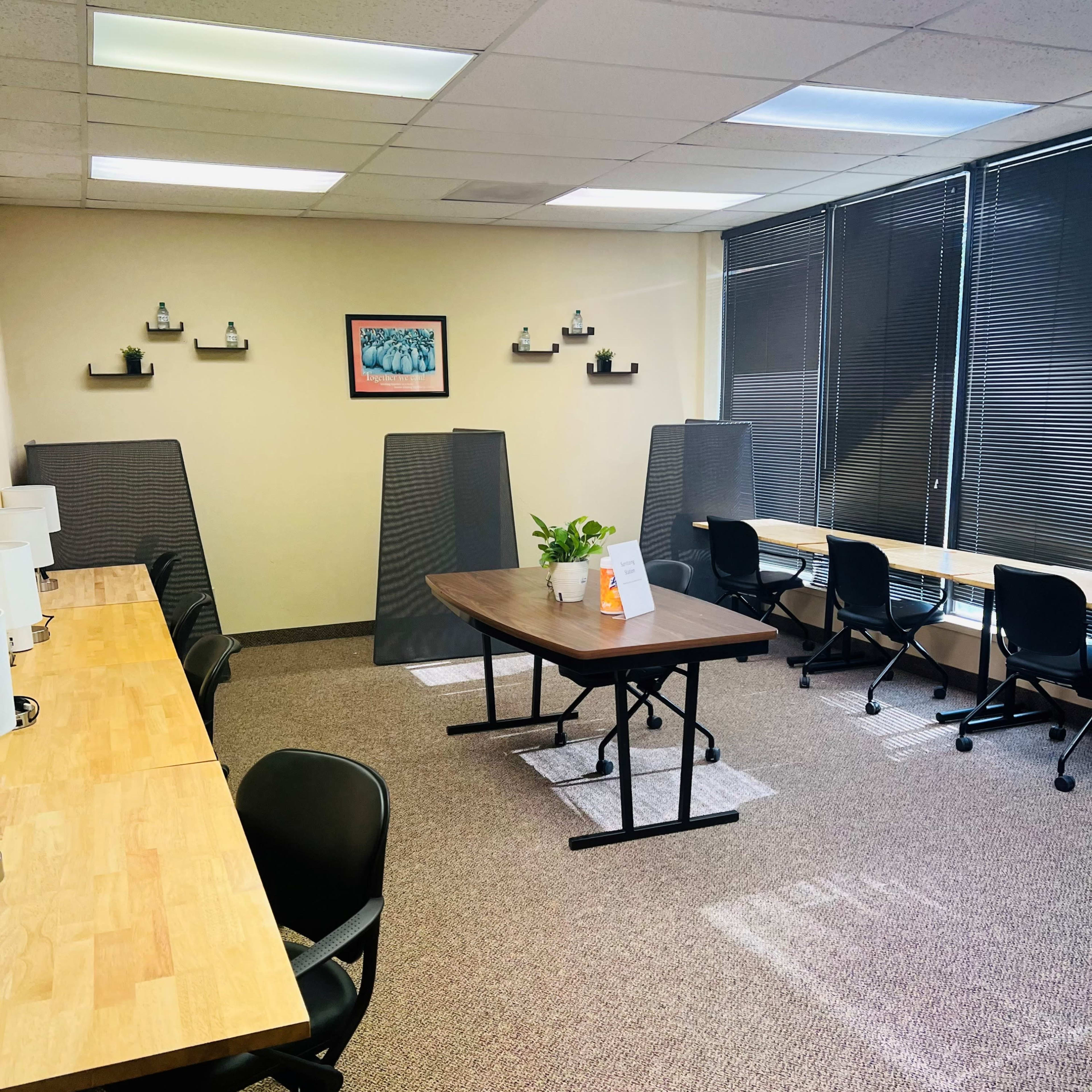 A modern office space with several workstations, a central table, and natural light coming through large windows.