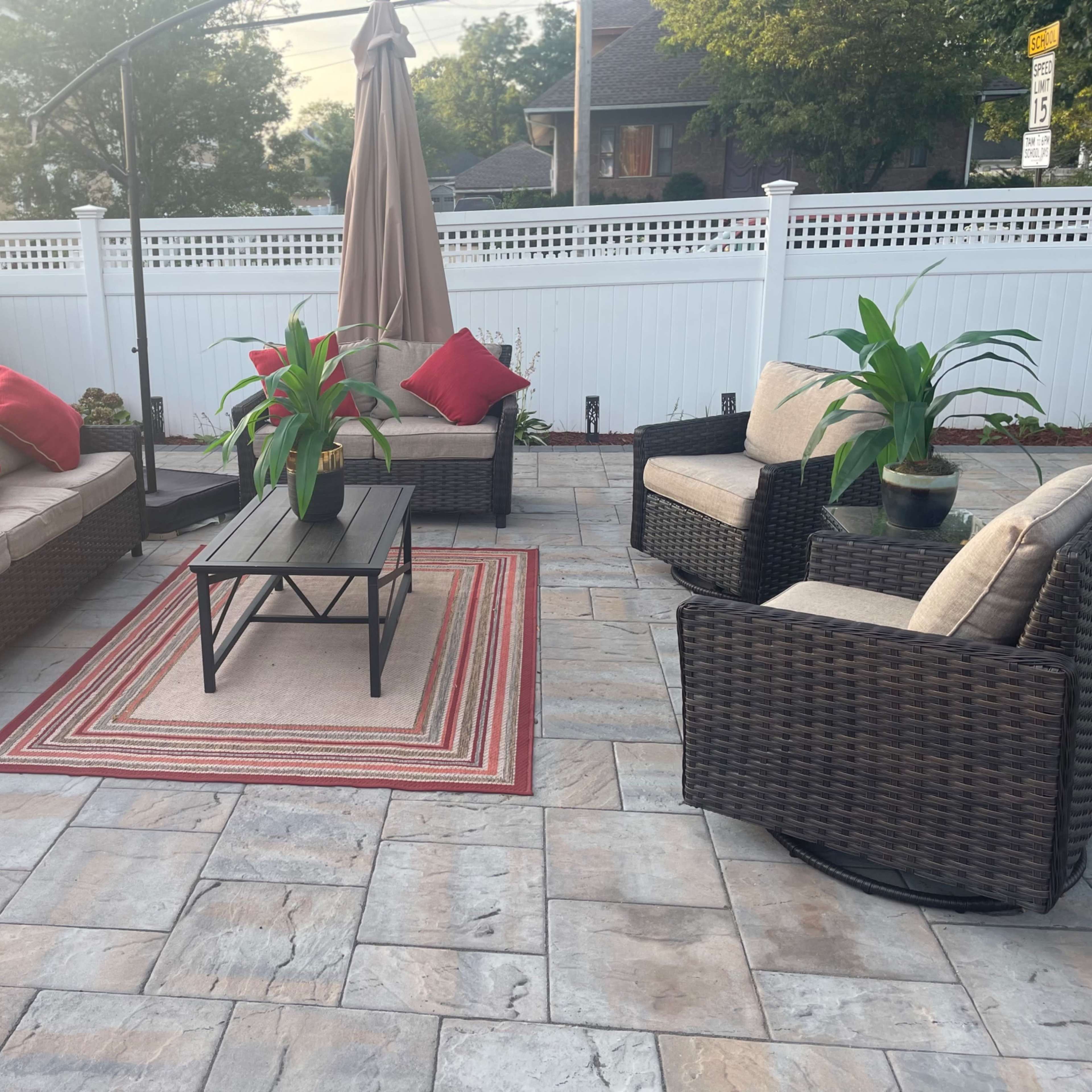 A patio area features a seating arrangement with beige cushions and red accent pillows around a central table, surrounded by potted plants and a decorative umbrella.