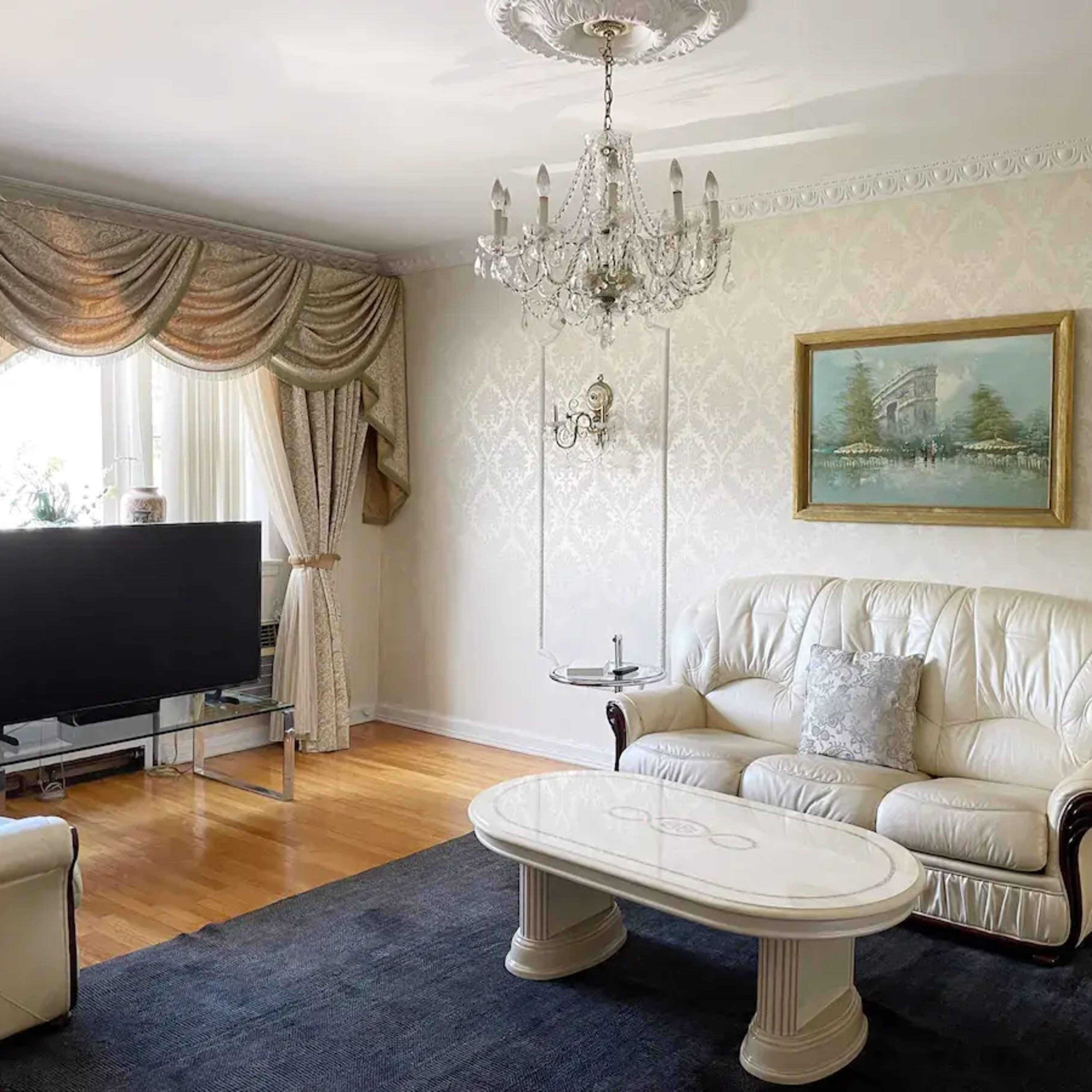 A living room features white leather sofas, a chandelier, a coffee table, and a large television against a patterned wall with an artwork above it.