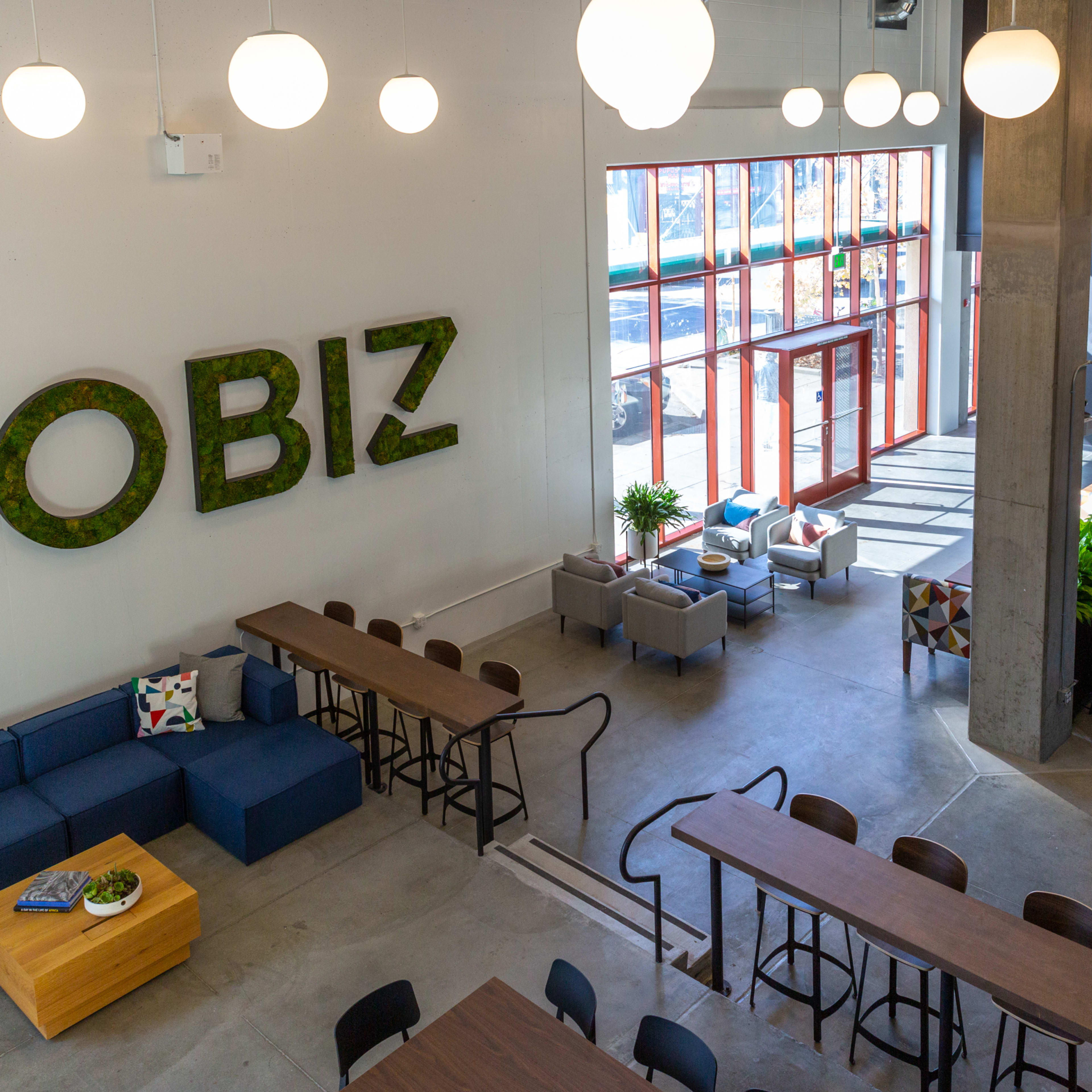 A spacious interior of a coworking space with large windows, green plants, and wooden tables arranged for seating.