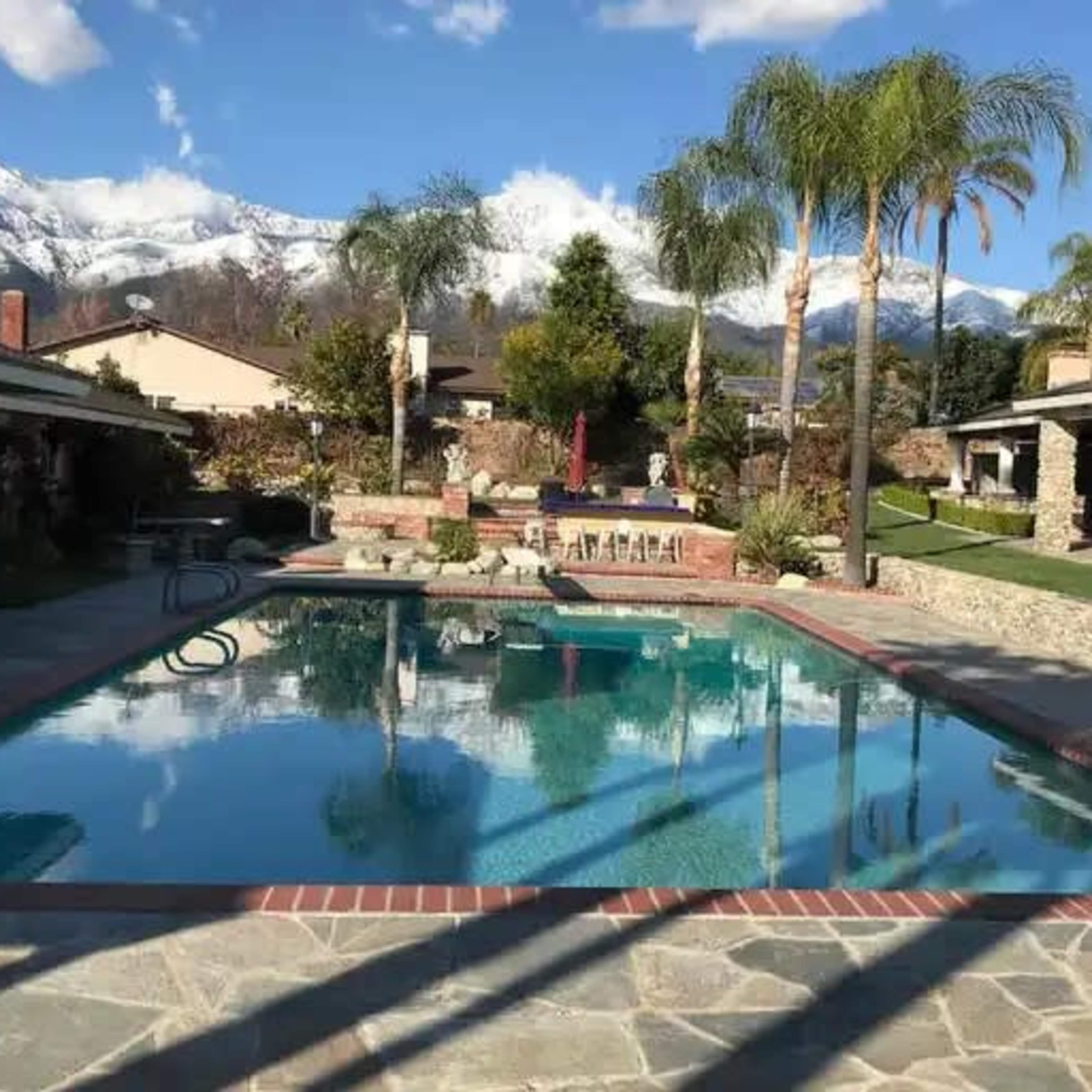 A clear swimming pool reflects the surrounding landscape, including palm trees and snow-capped mountains, in a landscaped backyard with several seating areas.