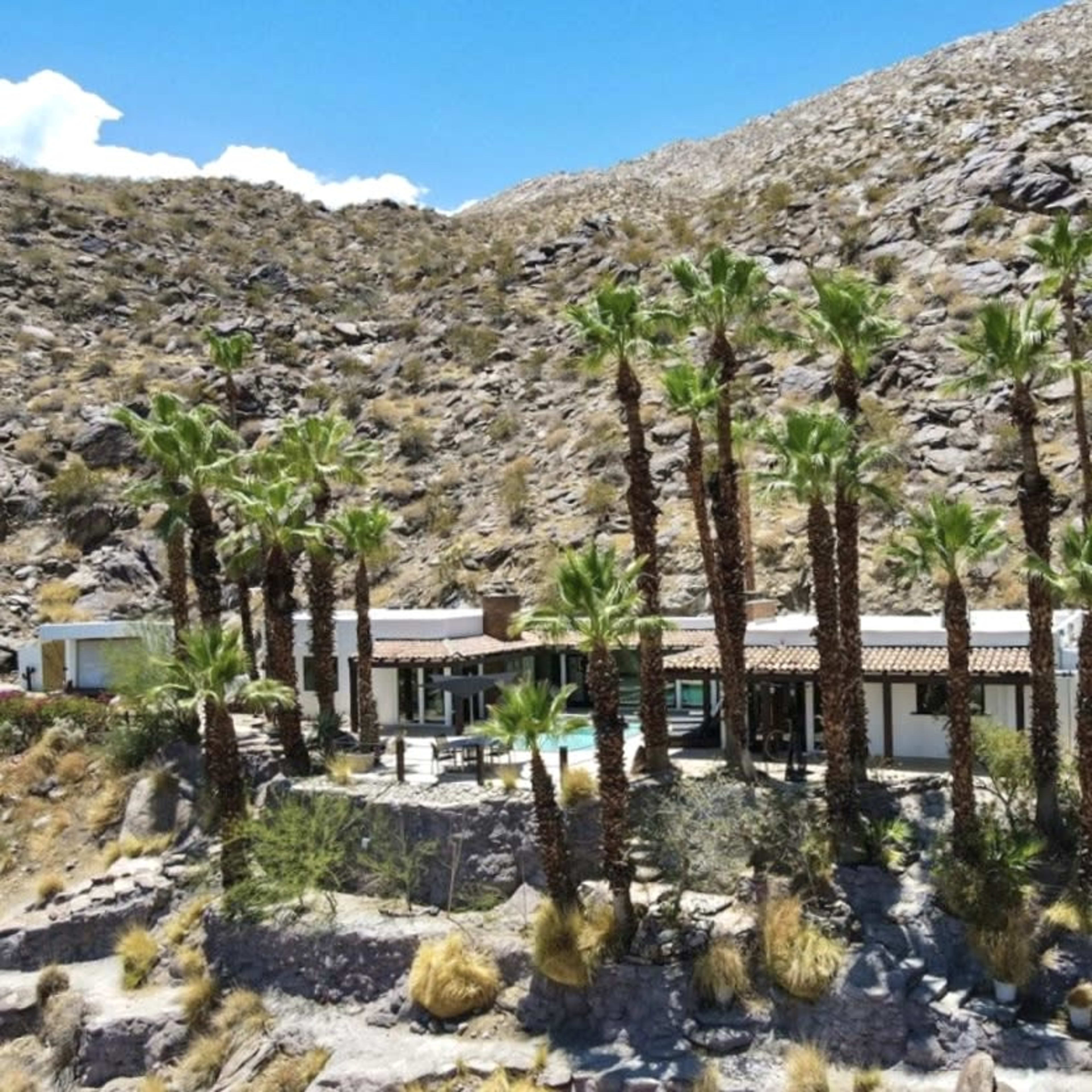 A modern house surrounded by tall palm trees, set against a mountainous, rocky landscape.