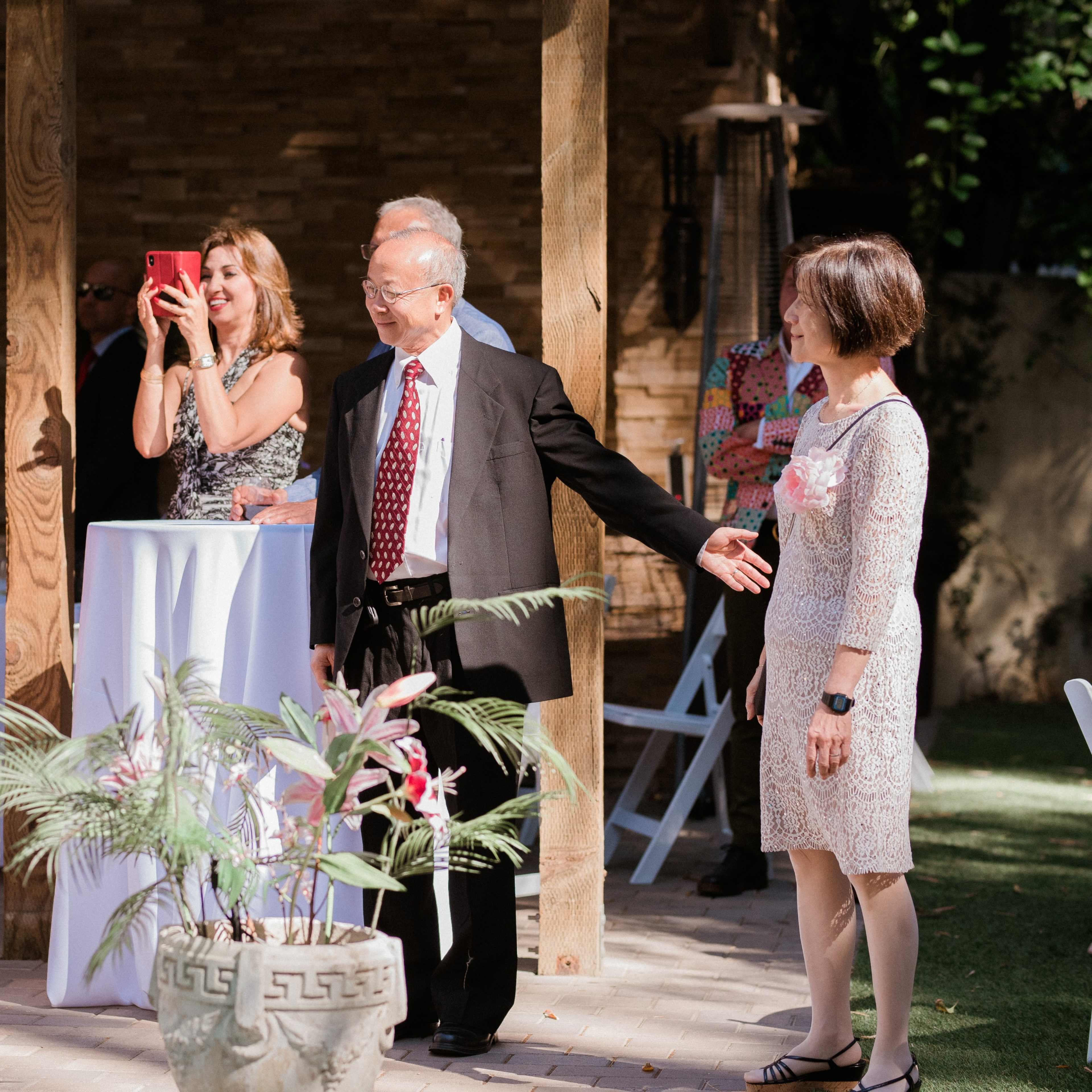 A bride wearing a white dress walks toward her family and friends, while one guest records the moment on a smartphone.
