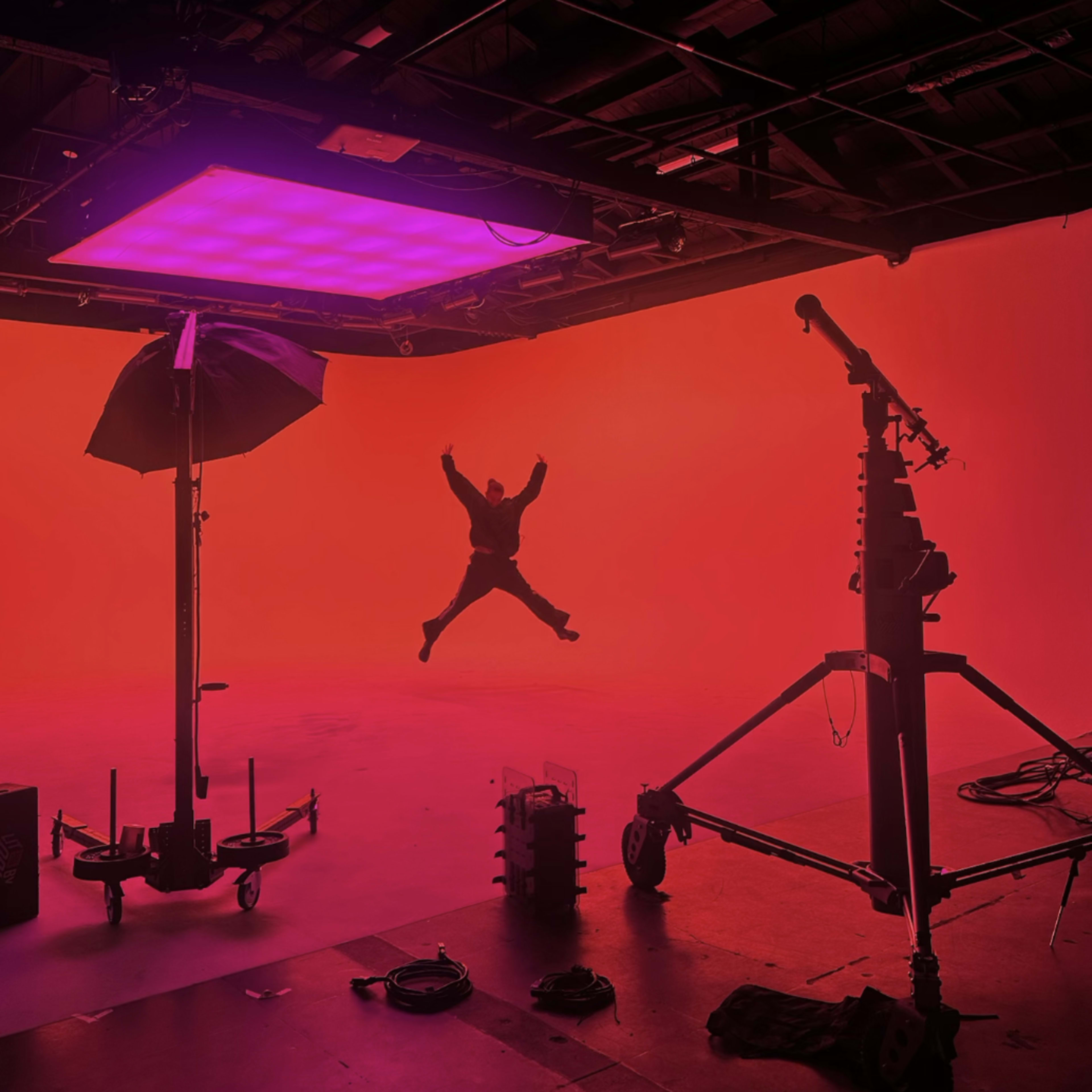 A person is jumping in a brightly lit studio with a vibrant orange background and lighting equipment setup.