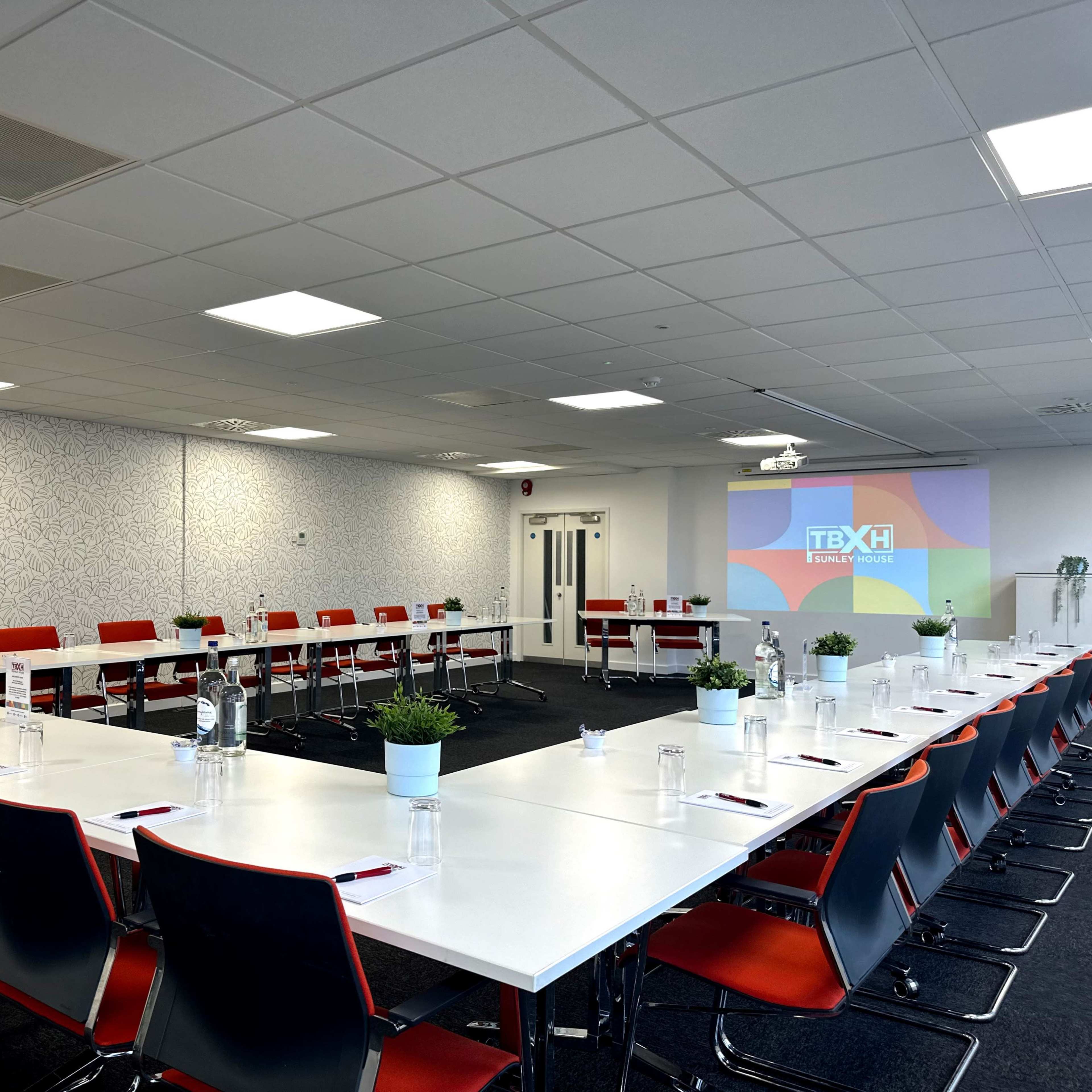 A spacious conference room features a large, U-shaped arrangement of white tables and red chairs, with a projector screen displaying a logo.