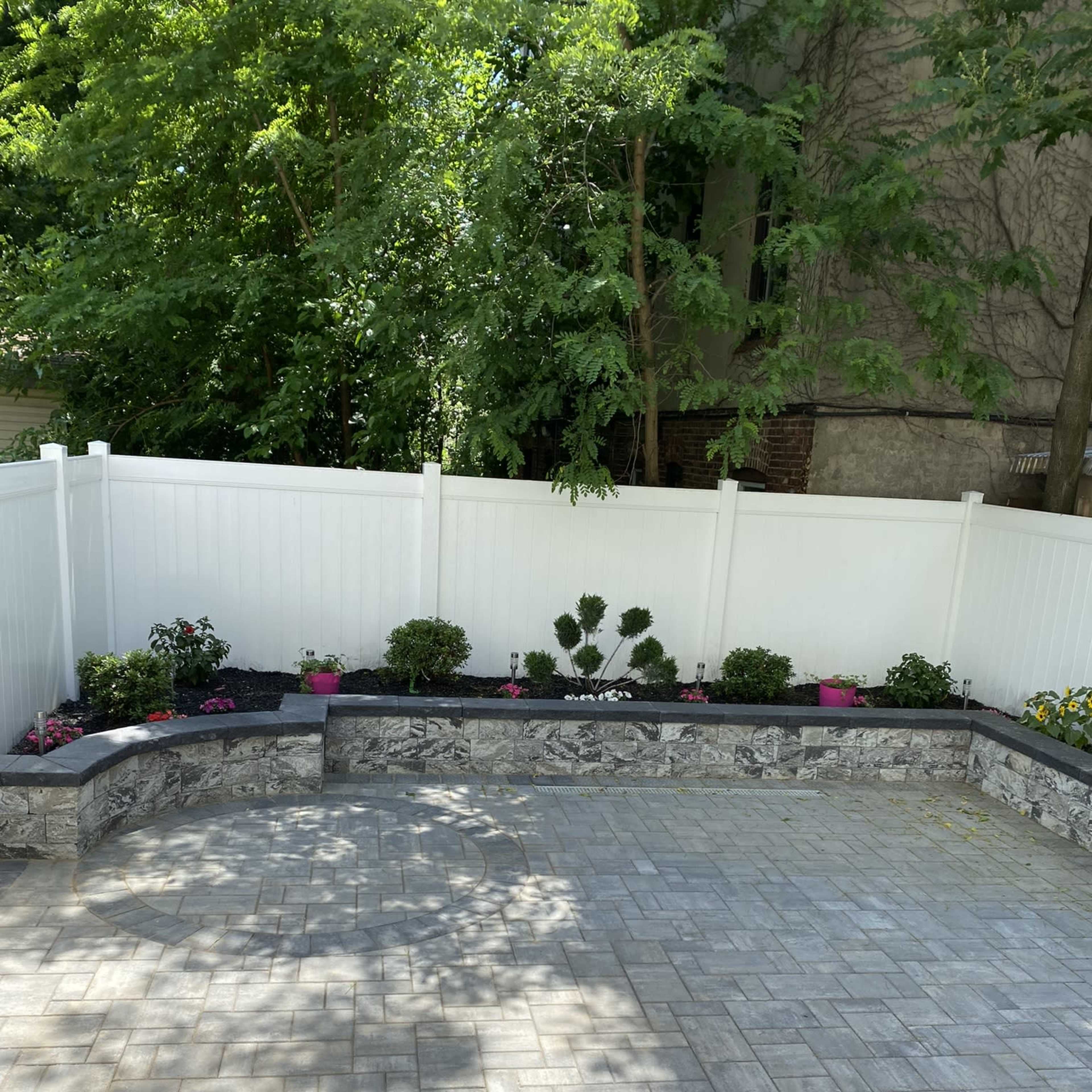A brick patio surrounded by a low stone wall with landscaped flower beds and a white fence in the background.