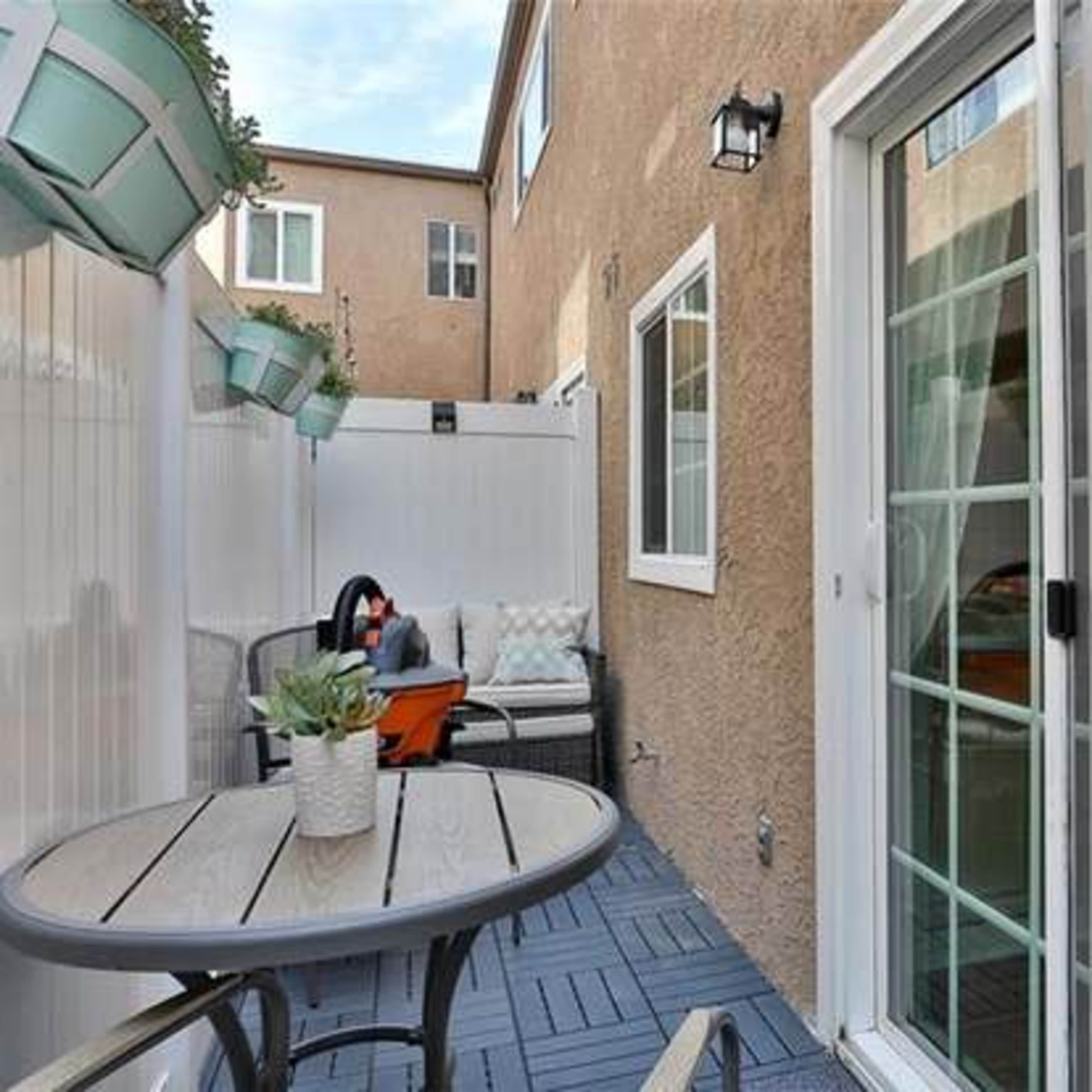 A small patio area features a round table and chairs, surrounded by a textured fence and plants in wall-mounted planters.