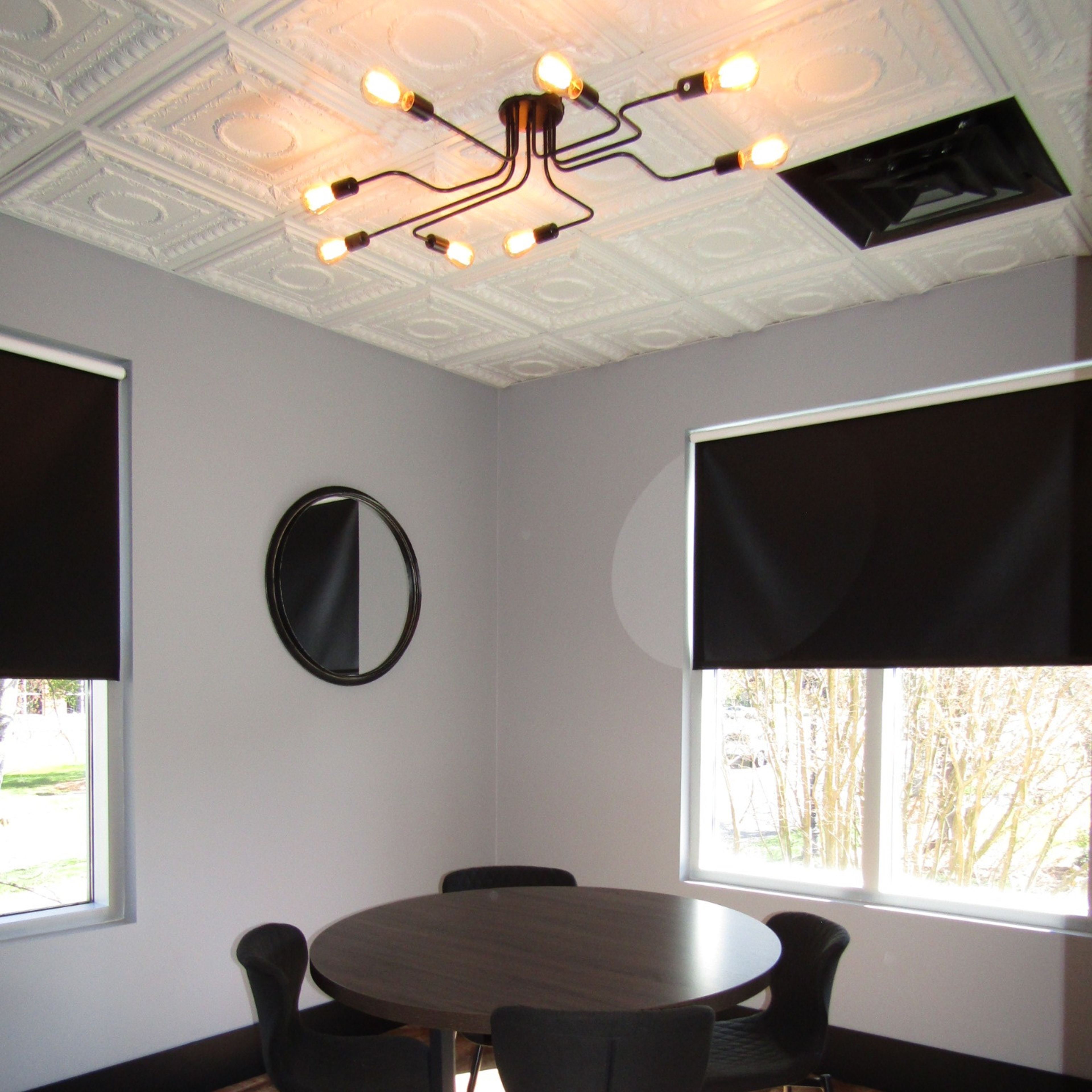 A dining area with a round table and four black chairs under a modern light fixture, featuring gray walls and large windows with black shades.