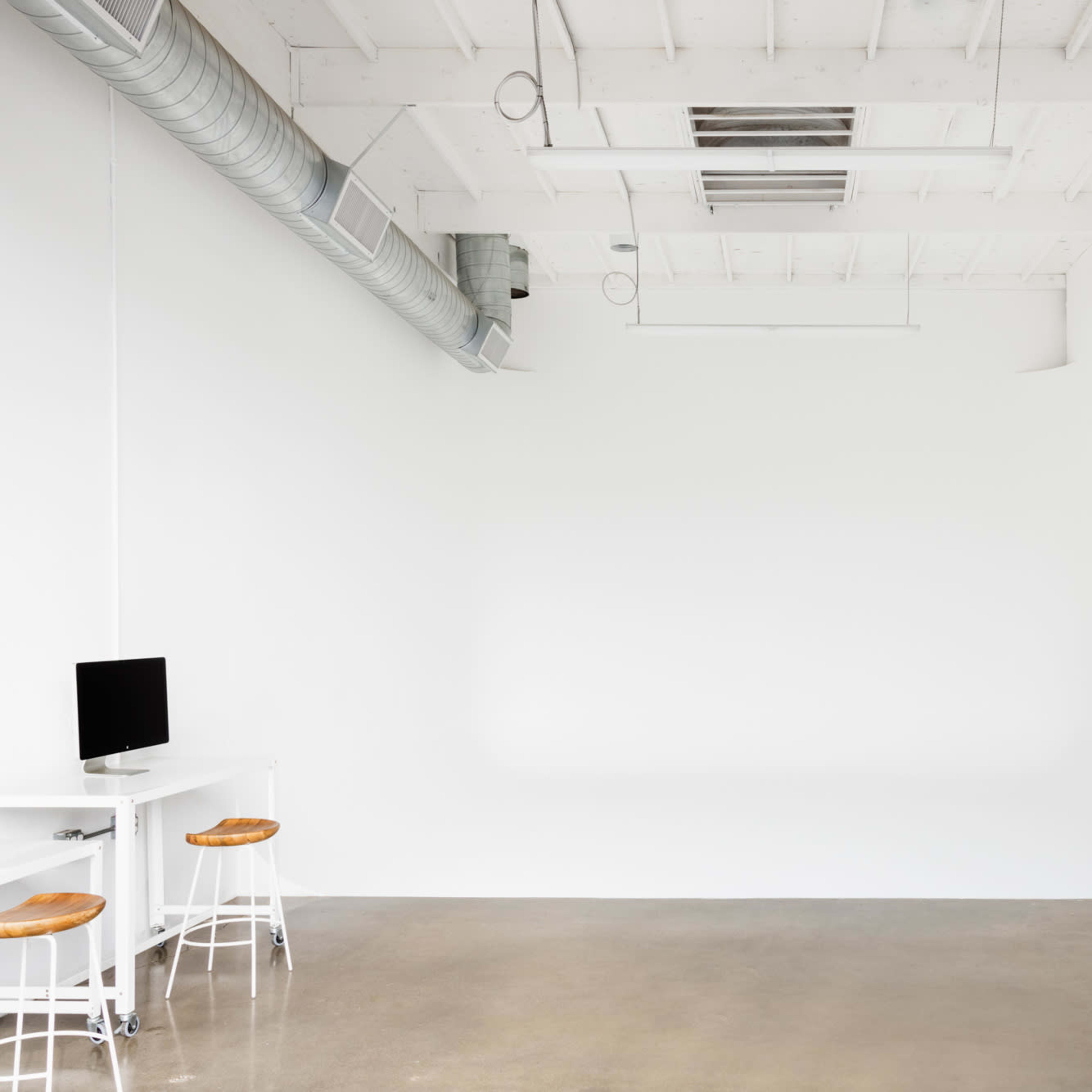 A minimalistic interior space with a polished concrete floor, white walls, and a few pieces of furniture, including a small desk with a computer and two stools.