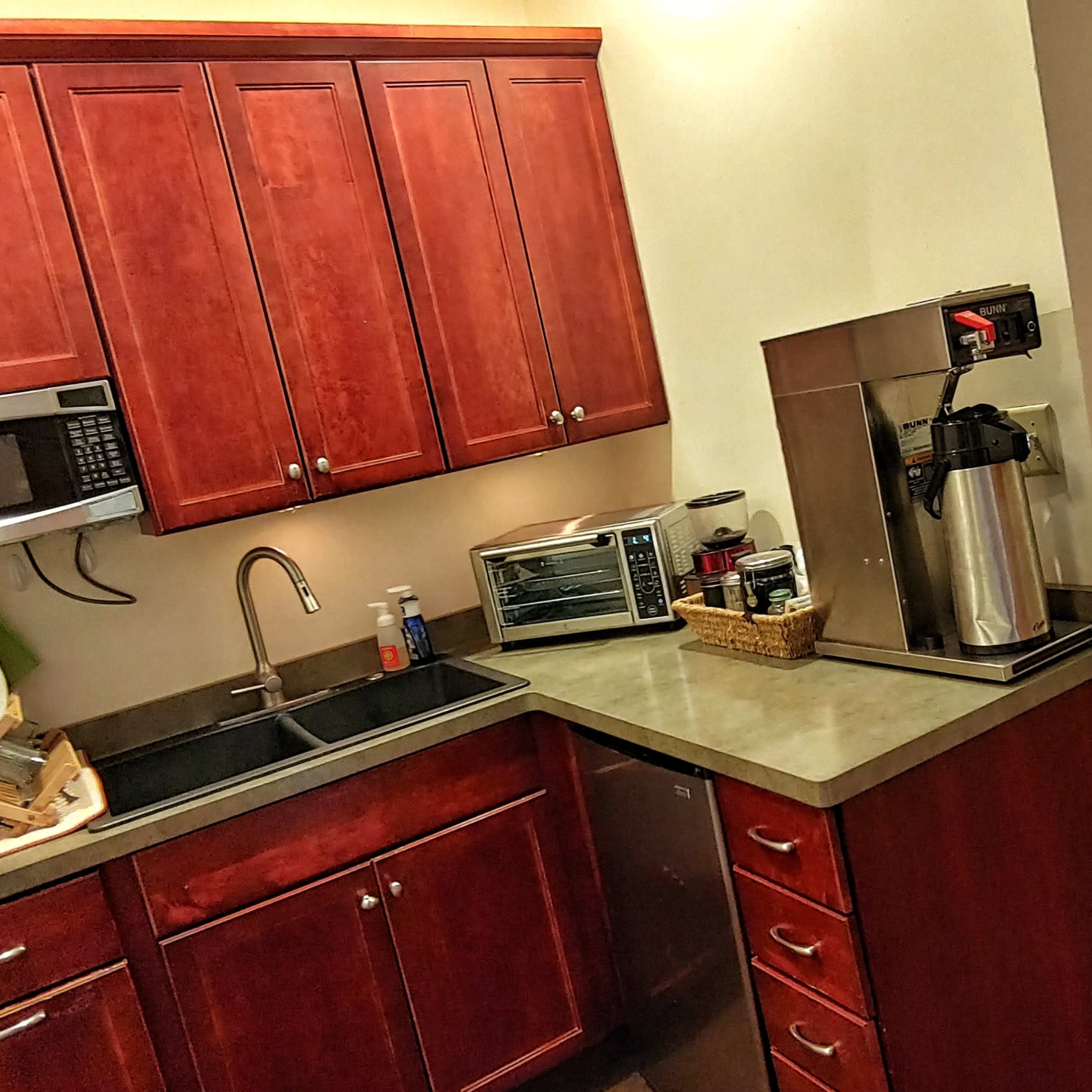 A kitchenette with wooden cabinets, a microwave, a sink, a countertop, and a coffee maker, adjacent to a hallway.