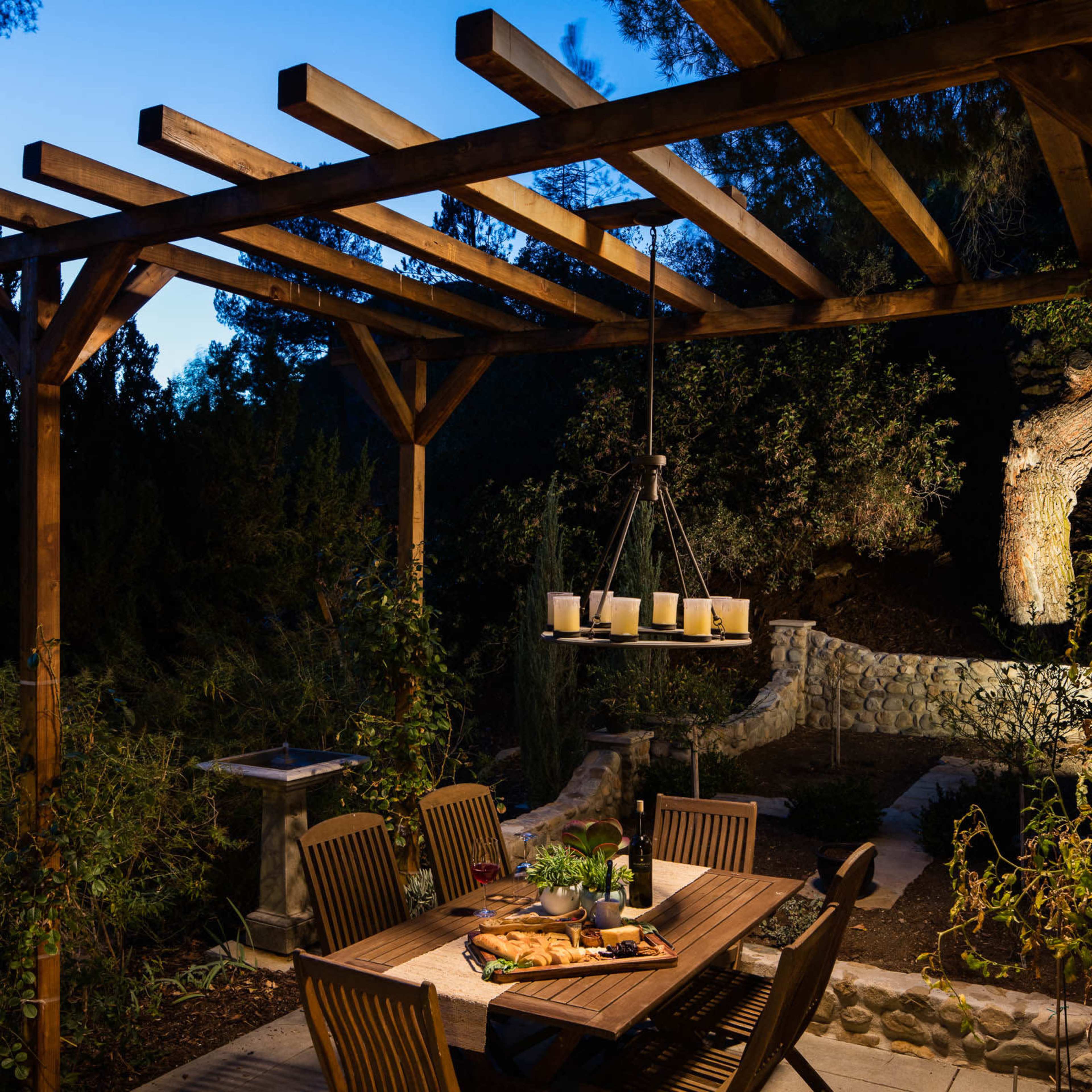 A wooden dining table is set with food and drinks under a pergola in an outdoor garden at dusk.