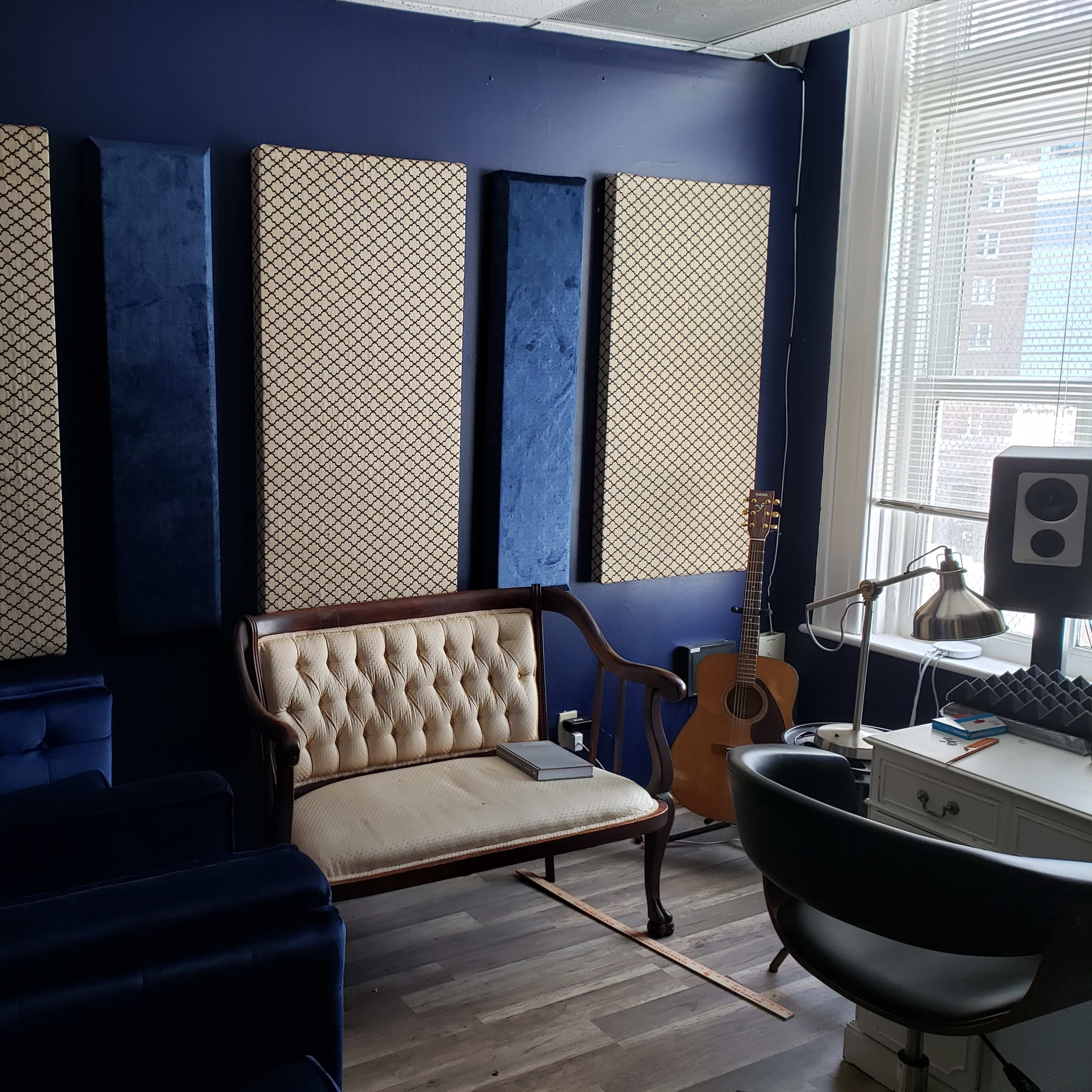 A cozy room with a blue and beige color scheme, featuring padded wall panels, a vintage-style sofa, a guitar, and a desk setup with audio equipment.