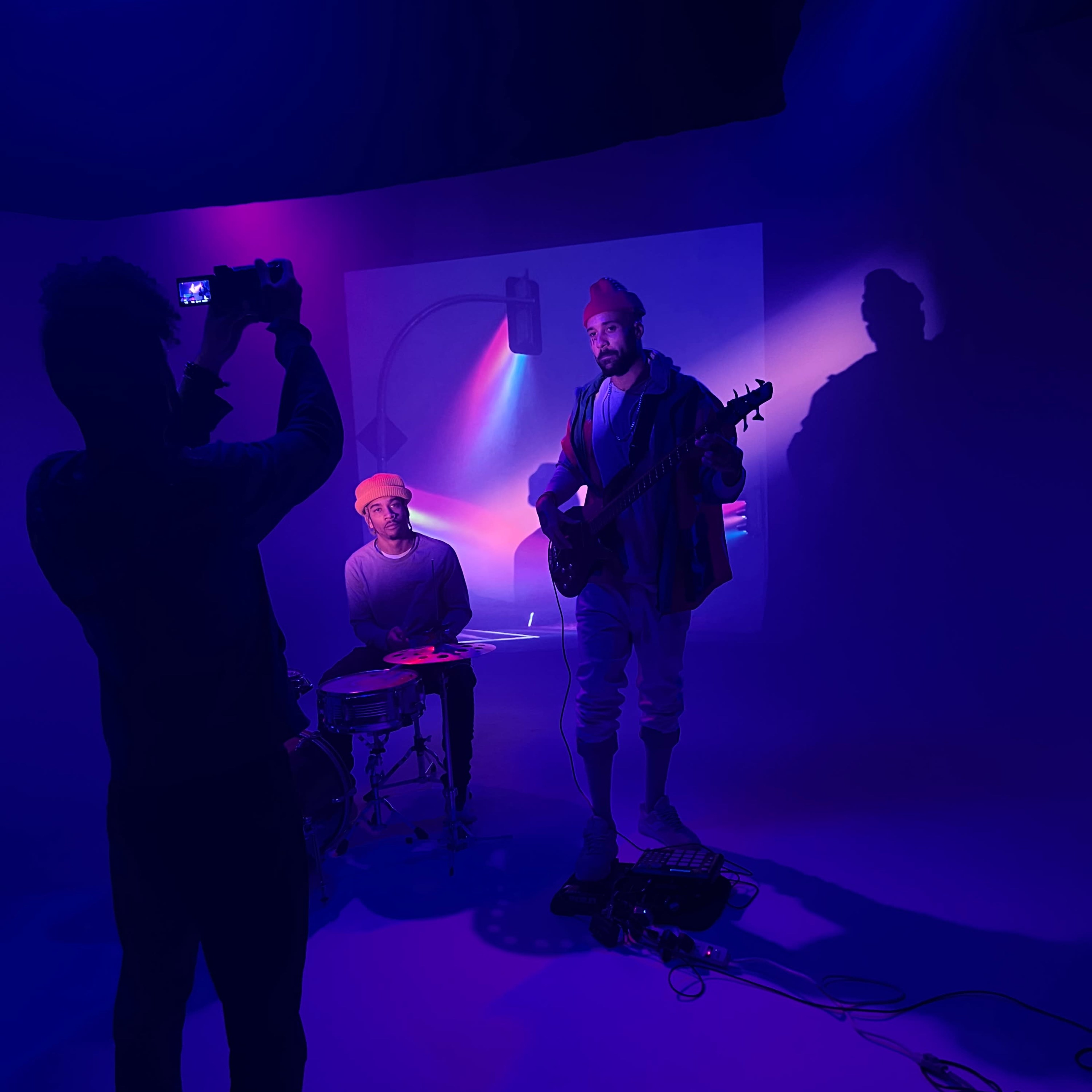 A musician plays guitar while another sits at a drum set, illuminated by colored lights in a studio setting.