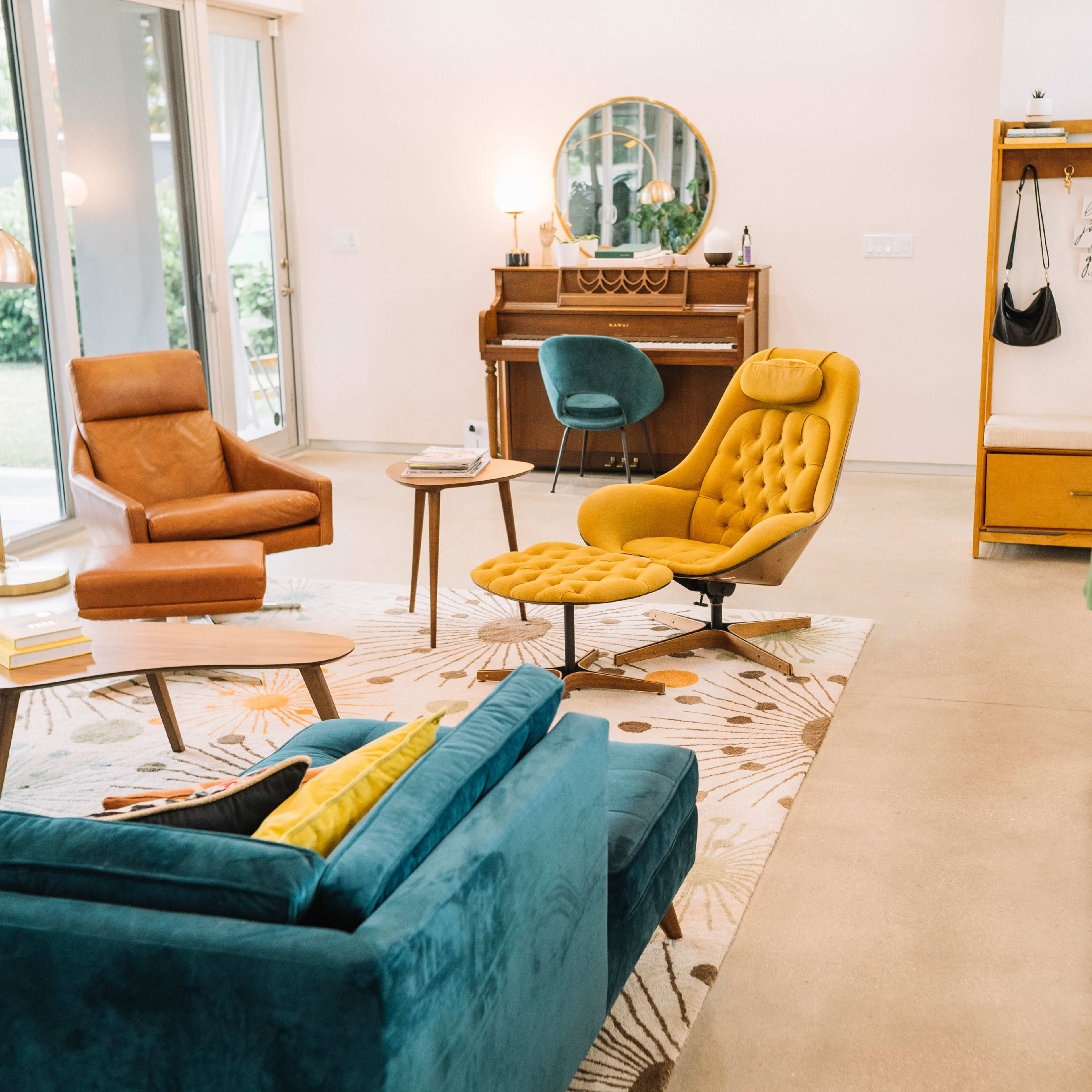 A modern living room features a blue couch, an orange chair, a yellow lounge chair, a round coffee table, and a piano, all arranged around a patterned rug.