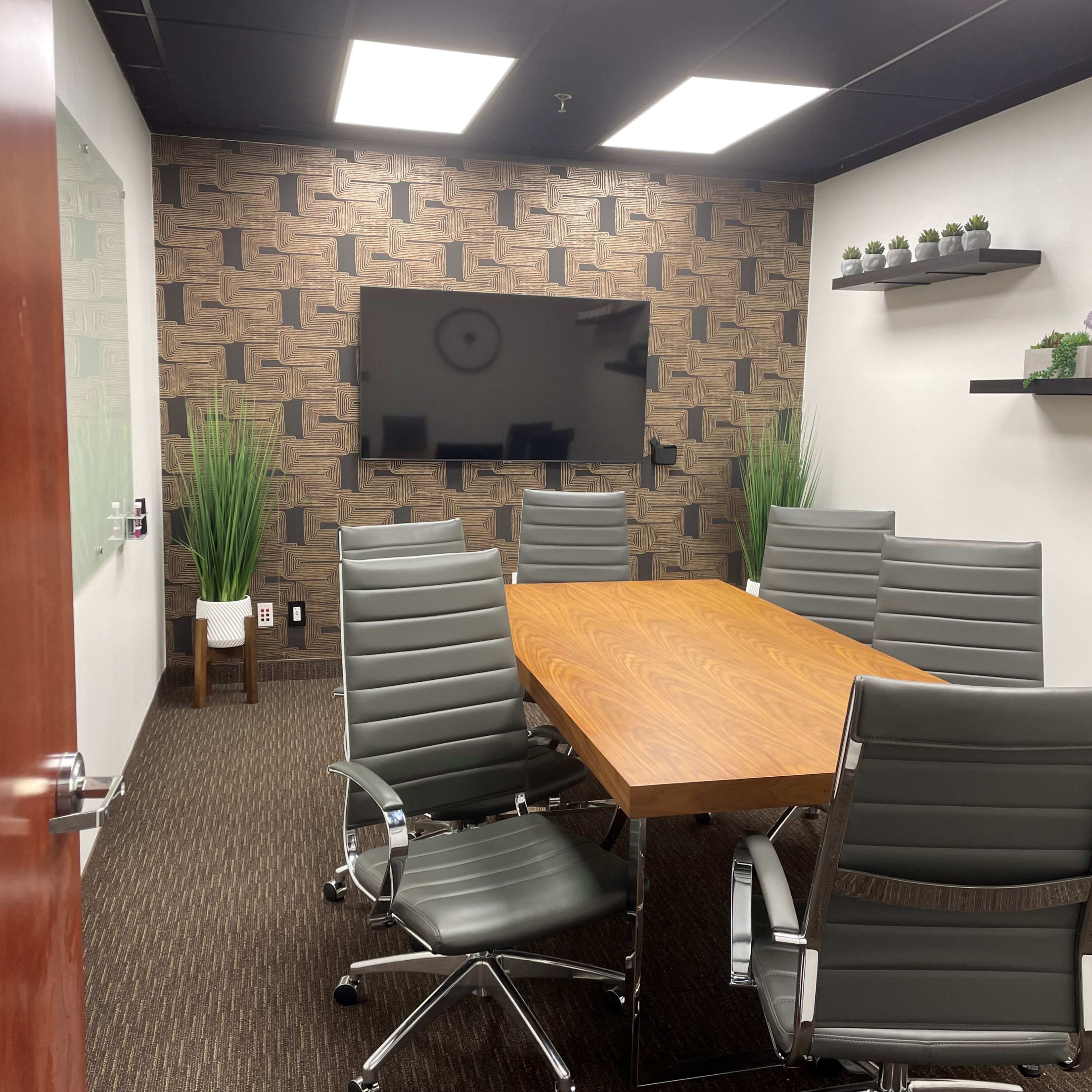 A conference room features a wooden table surrounded by six gray leather chairs, with a wall-mounted television and decorative plants on shelves.