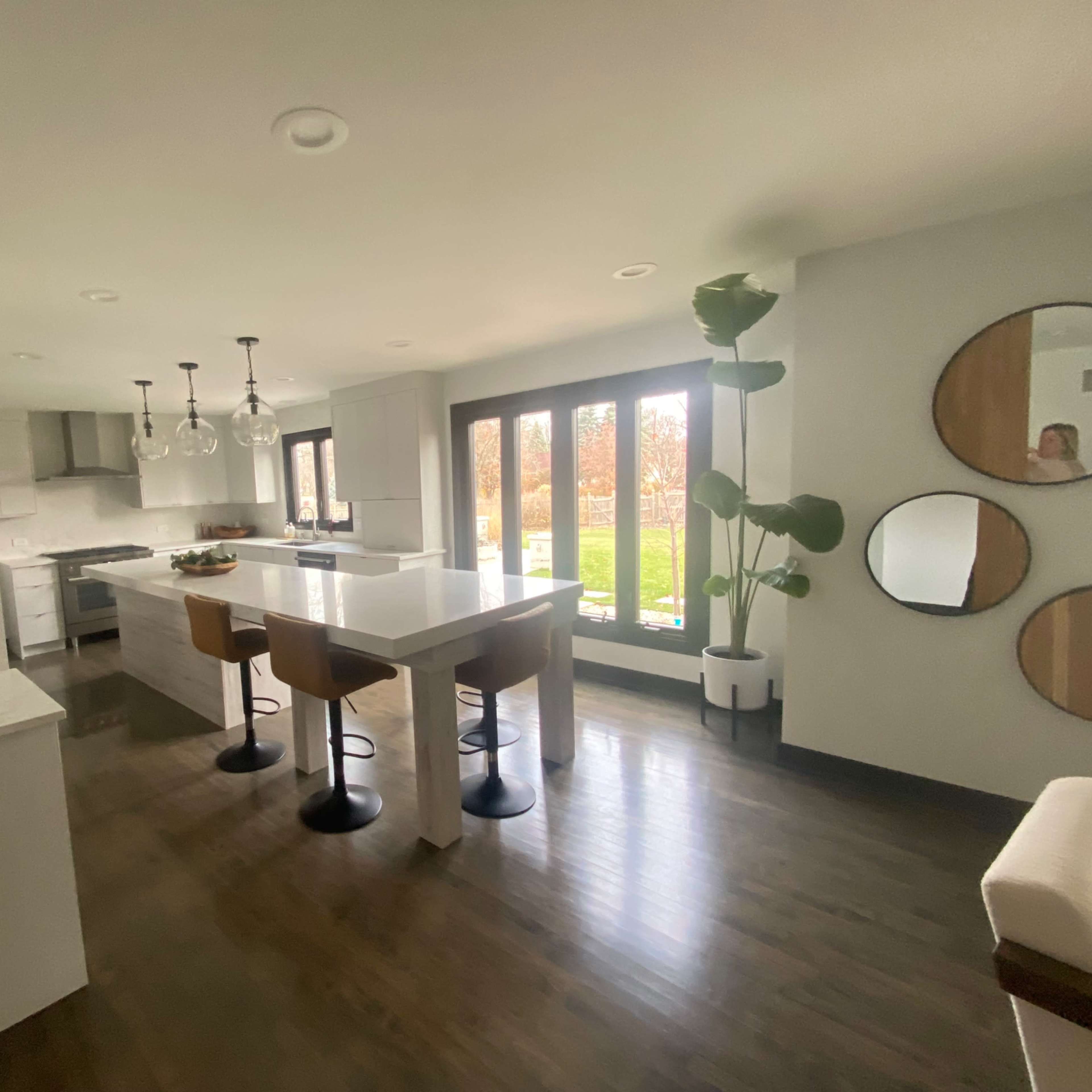 A modern kitchen and dining area with a central island, four bar stools, and large windows overlooking a garden.
