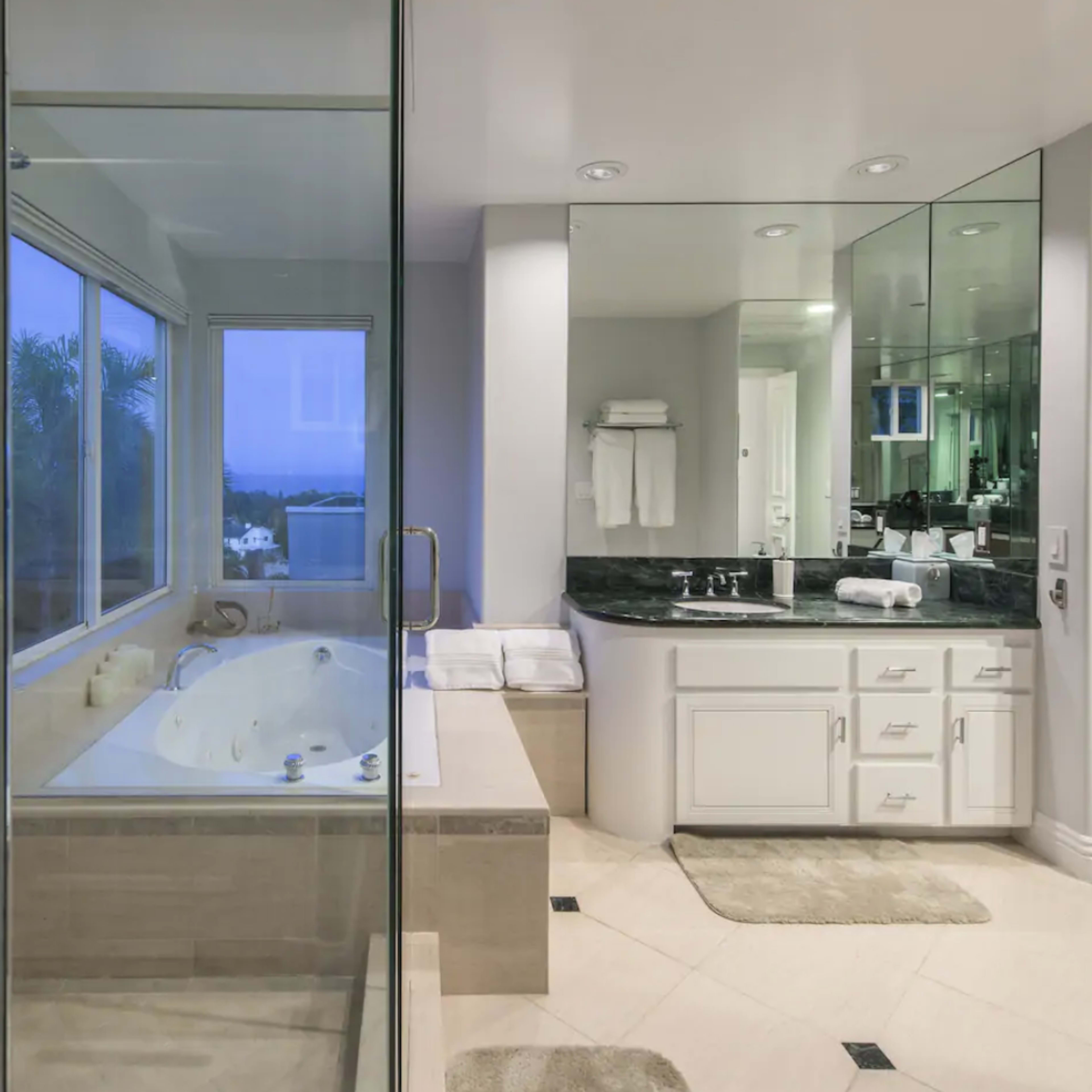 A modern bathroom featuring a glass-enclosed shower, a soaking tub by a large window, and a double vanity with a dark countertop.