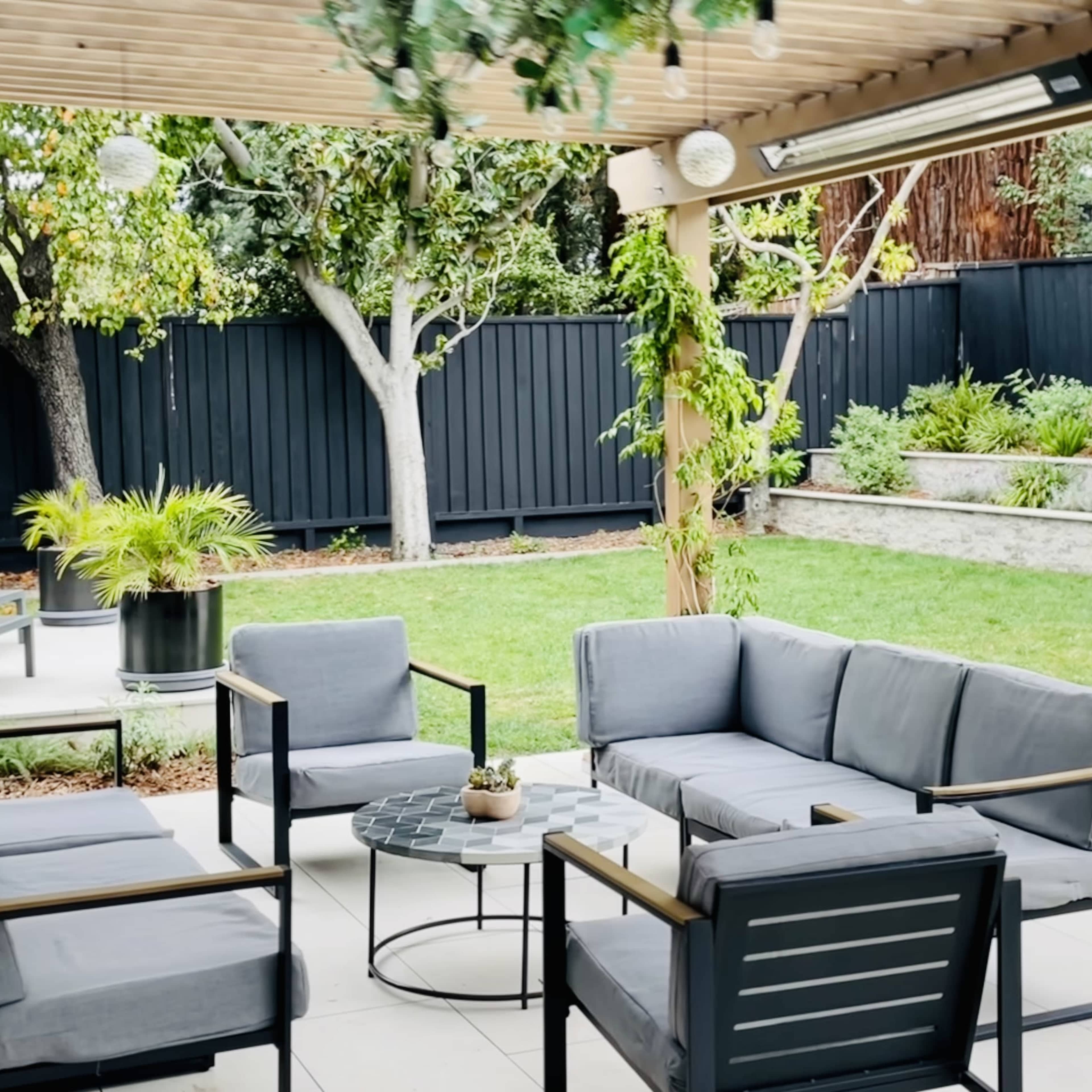 A modern outdoor seating area features grey fabric chairs and a coffee table under a wooden pergola, surrounded by a manicured lawn and landscaped garden.