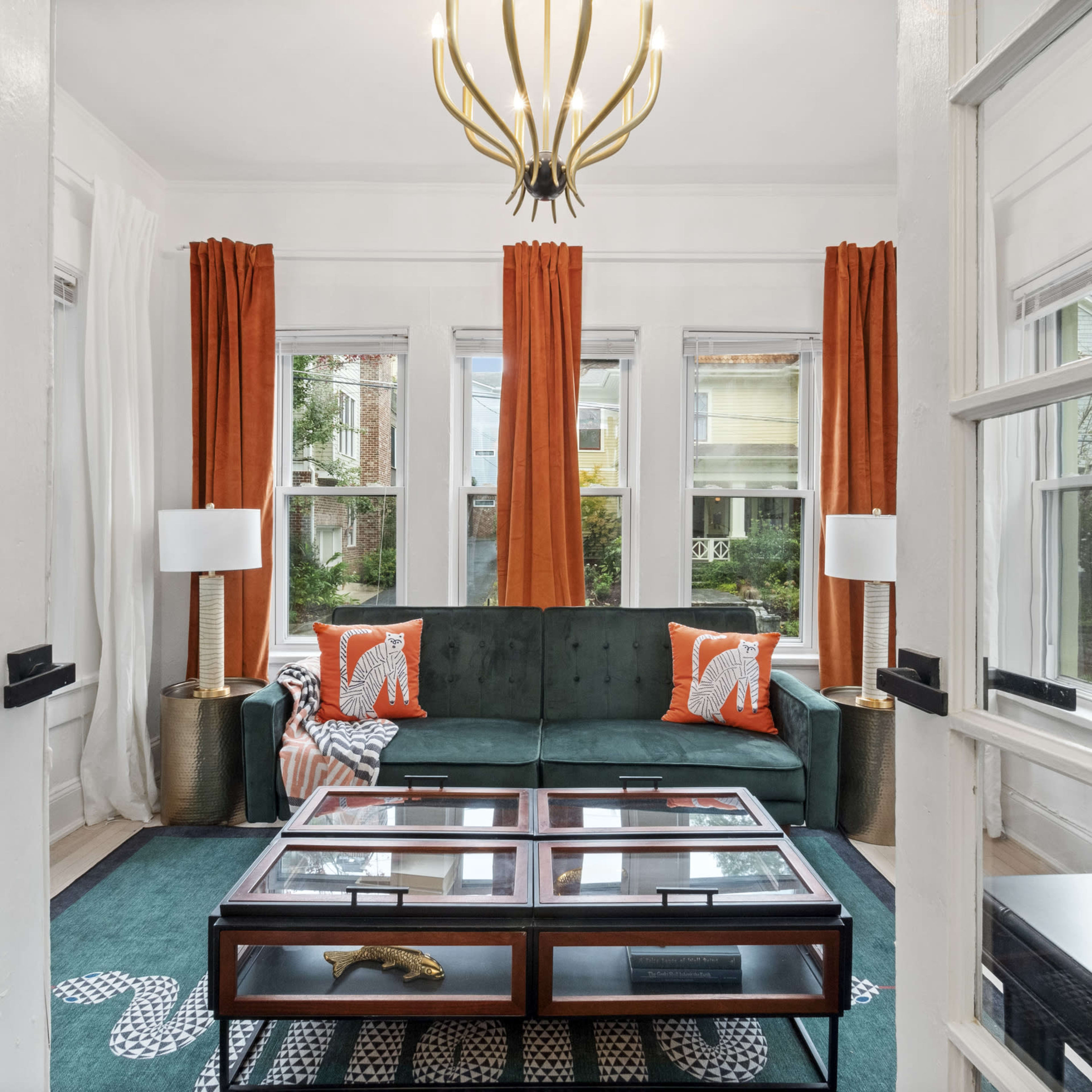 A bright living room featuring a green sofa, large windows with orange curtains, and a coffee table with a glass top.