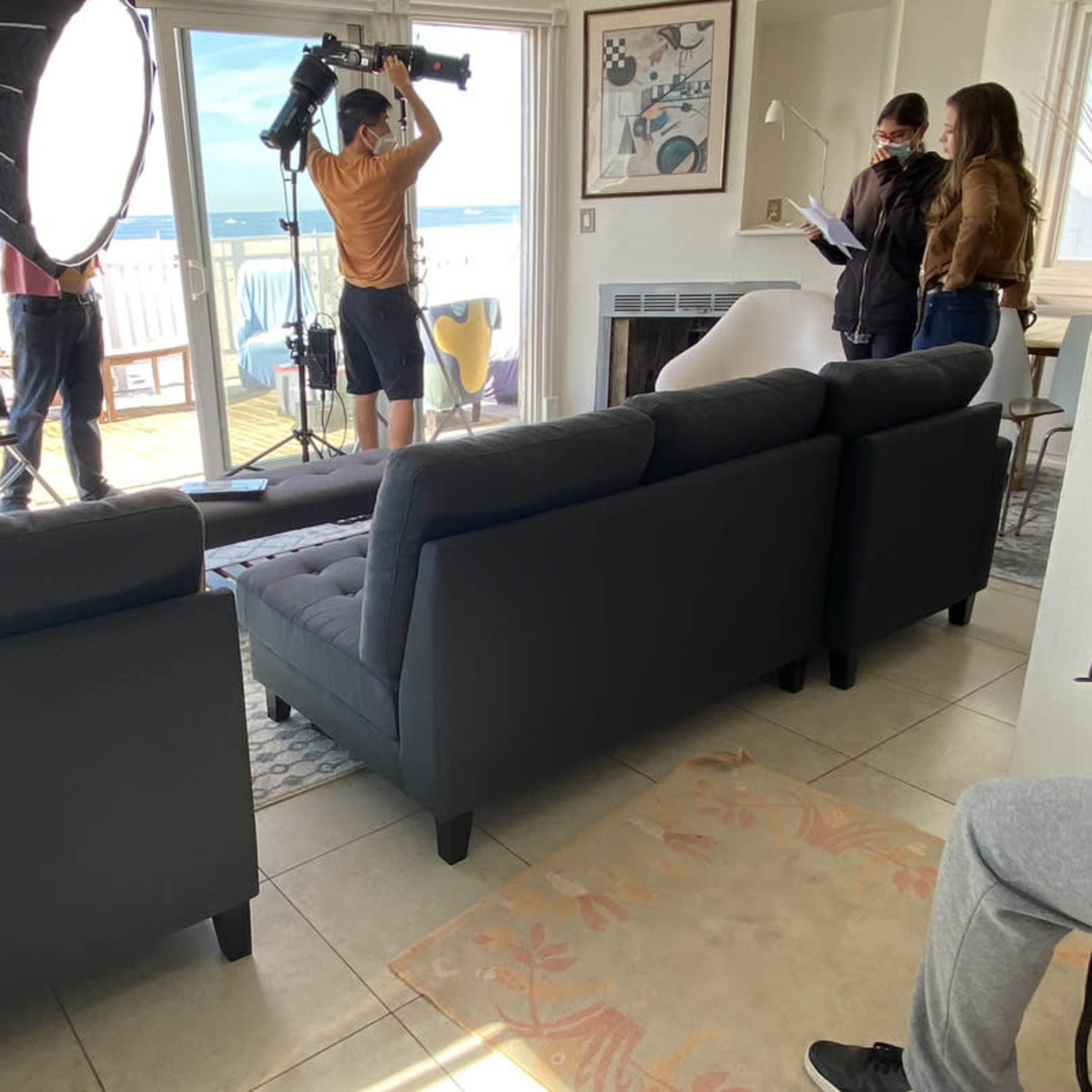 A film crew sets up equipment in a living room overlooking a beach.