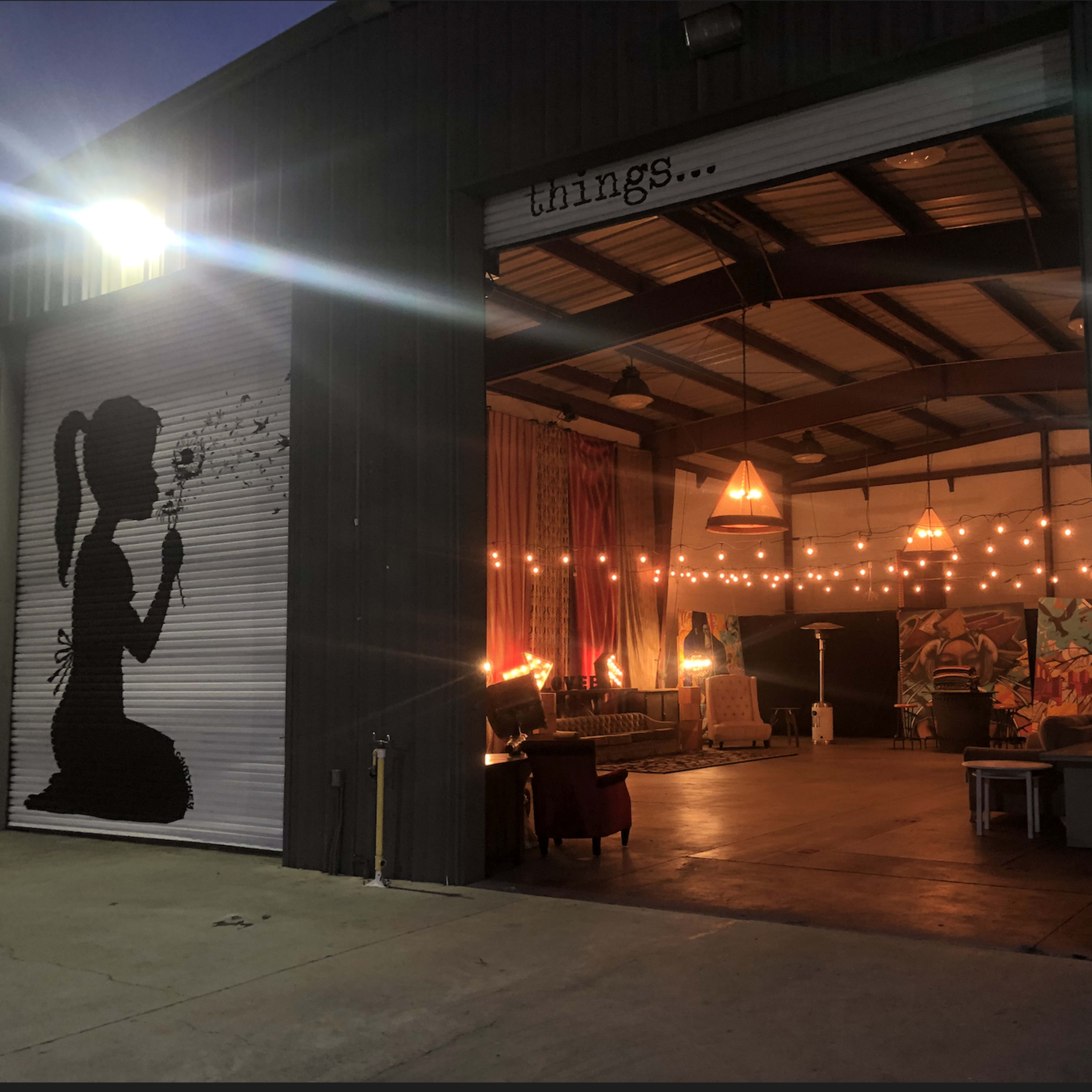 A large industrial garage door is open, revealing an indoor space decorated with lights, art, and furniture.