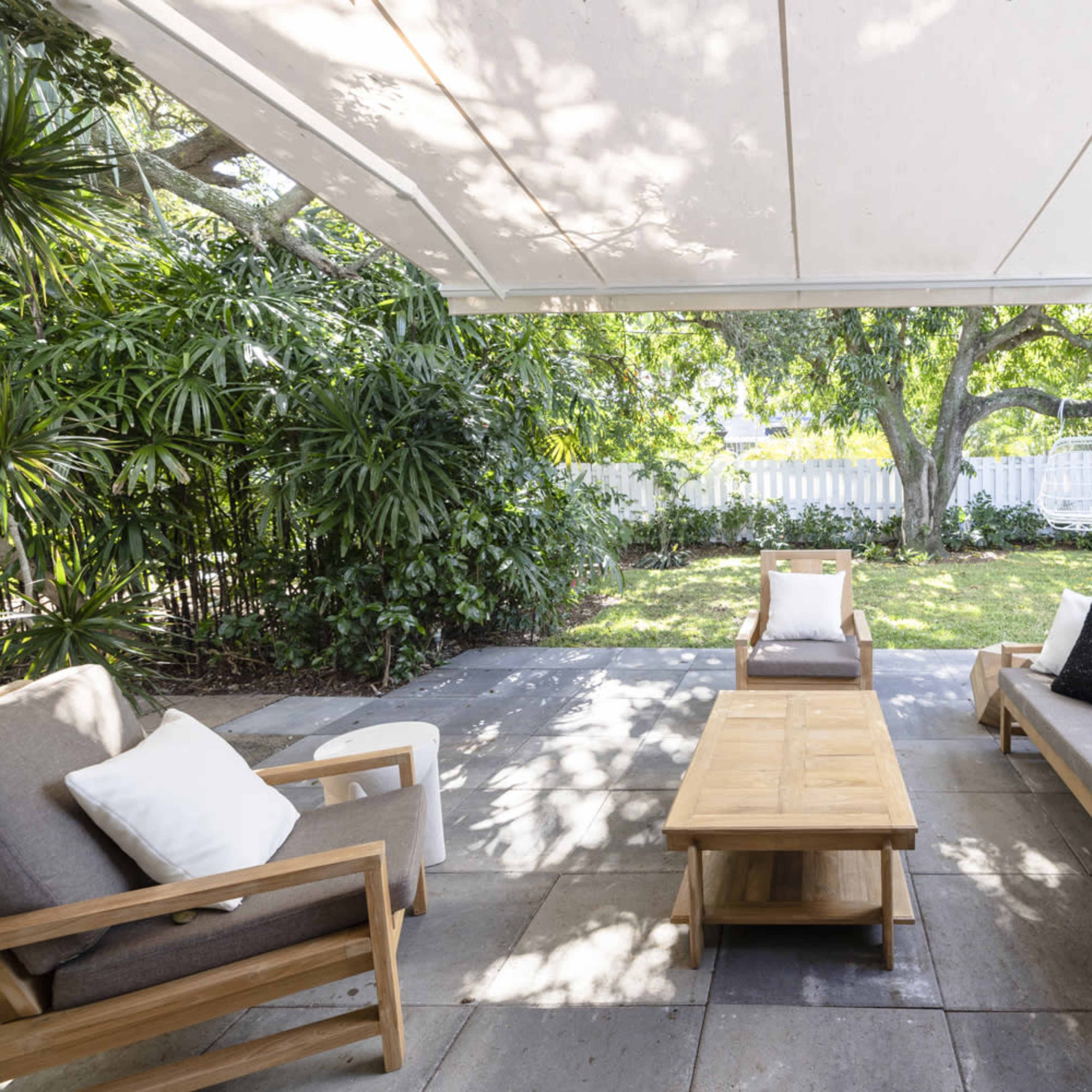 A covered patio features wooden furniture, including a coffee table and two chairs, surrounded by greenery and a lawn.