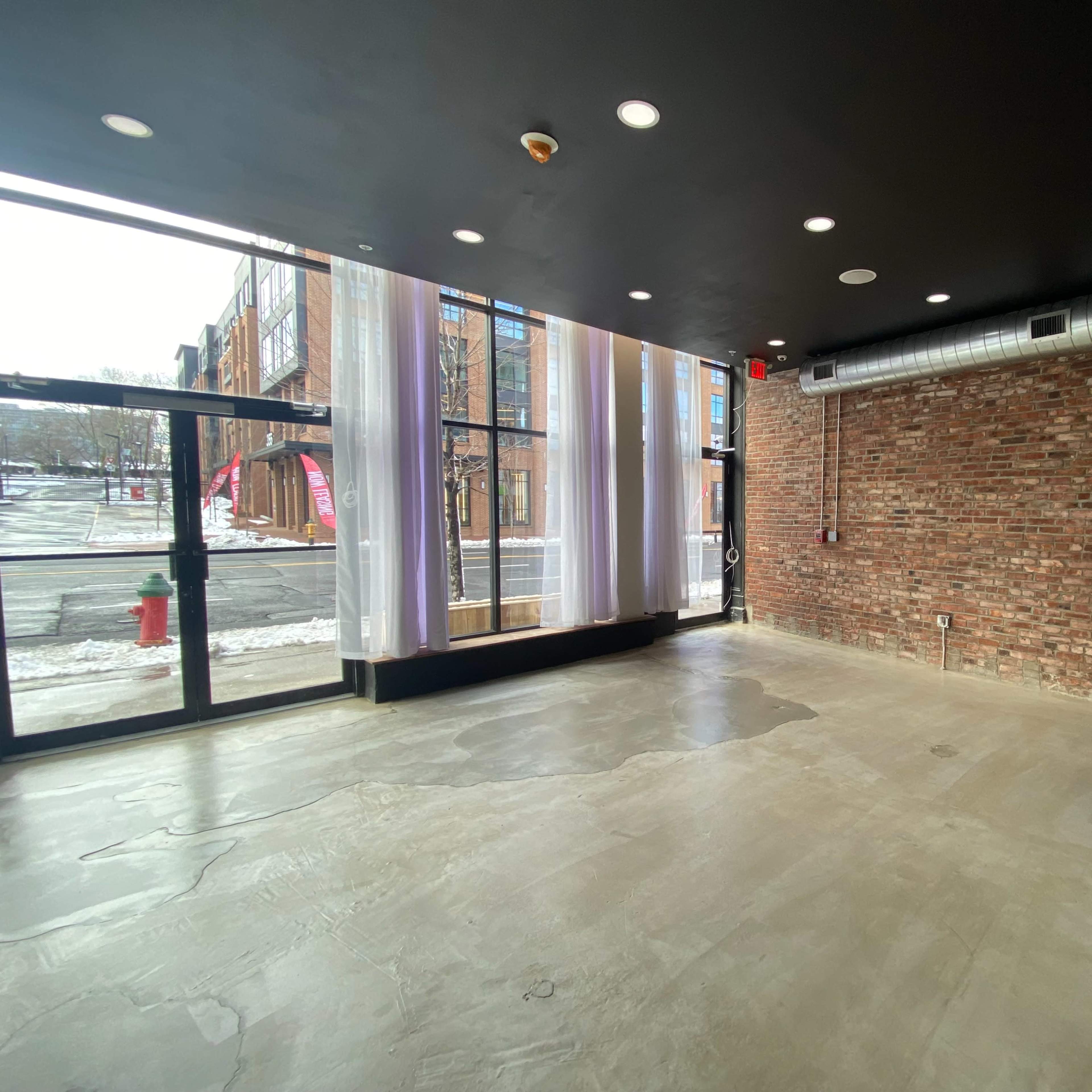 A vacant commercial space with exposed brick walls, large windows, and a concrete floor.