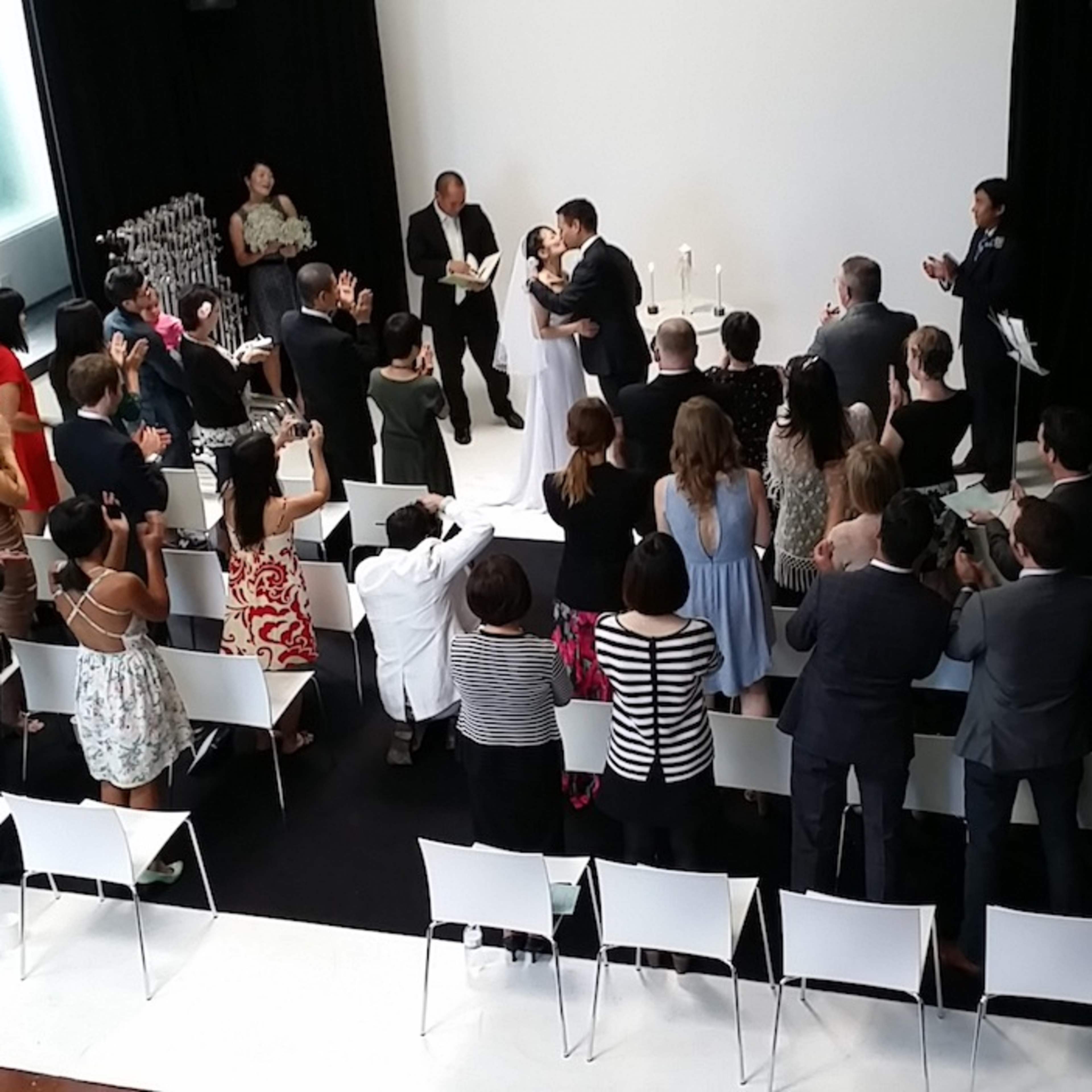 A wedding ceremony is taking place in a minimalist indoor venue, with guests seated and standing around the couple at the front.