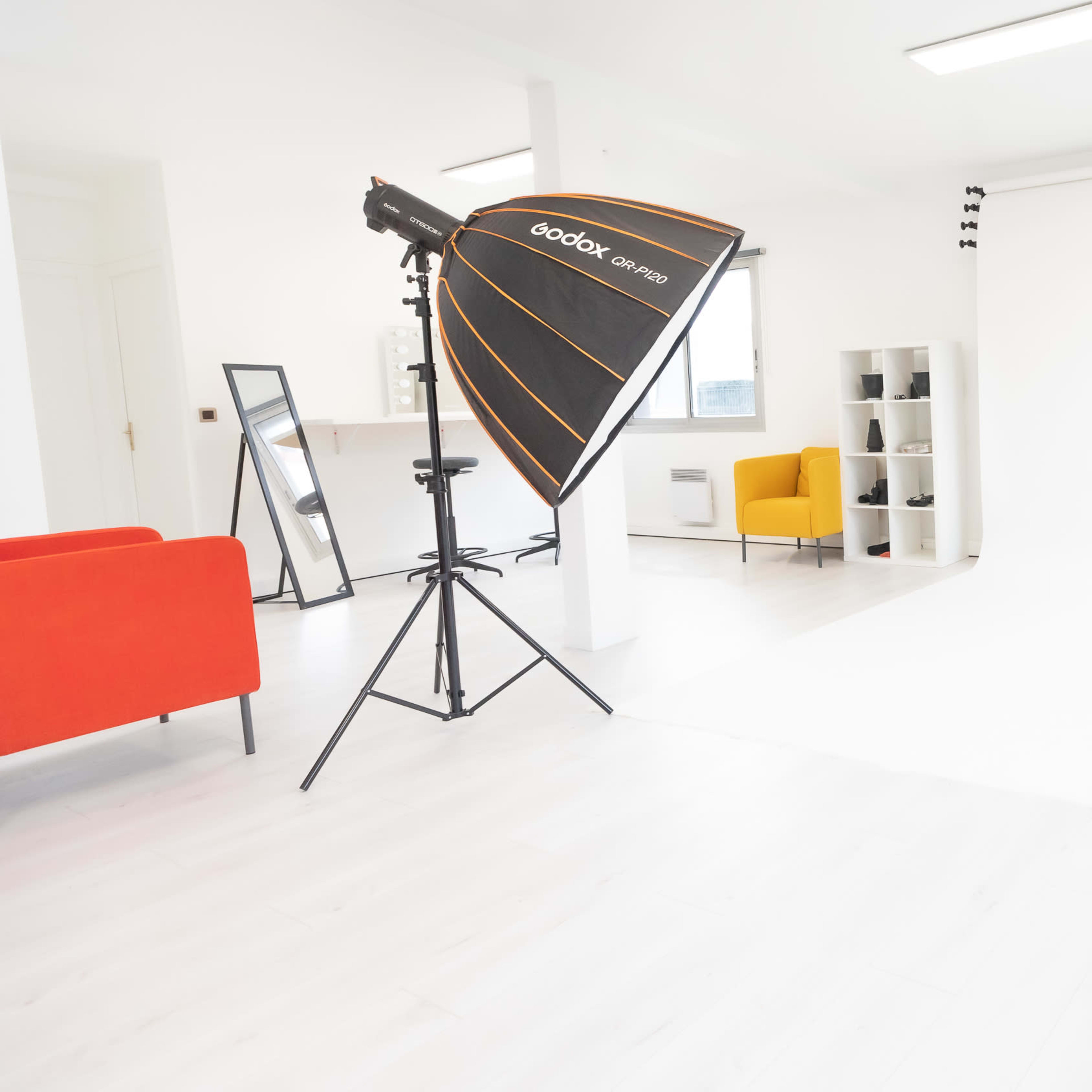A well-lit photography studio featuring a white background, softbox lights on stands, a yellow chair, and a red couch.