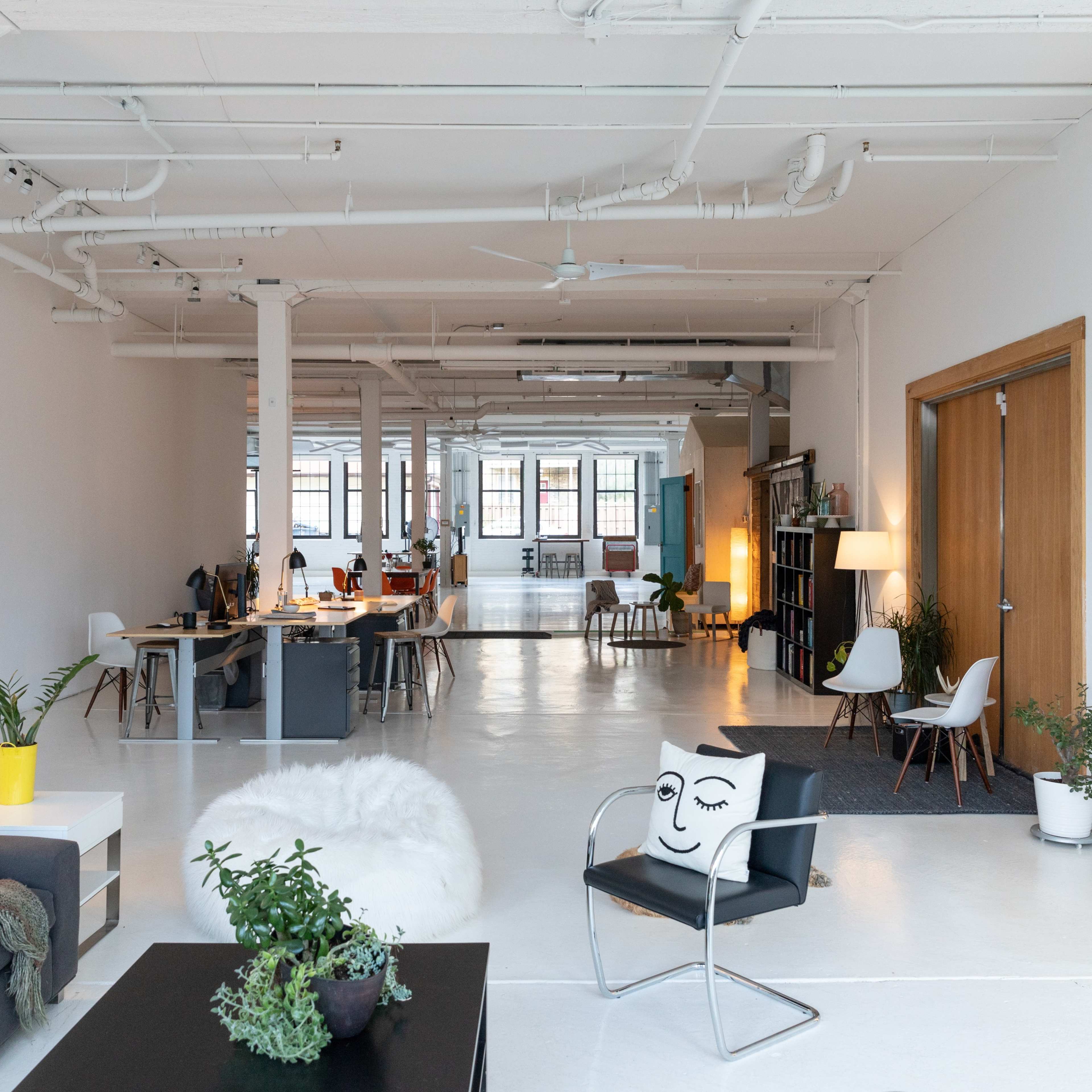 A spacious, modern office area with desks, seating, and plants arranged in a bright, minimally decorated interior.