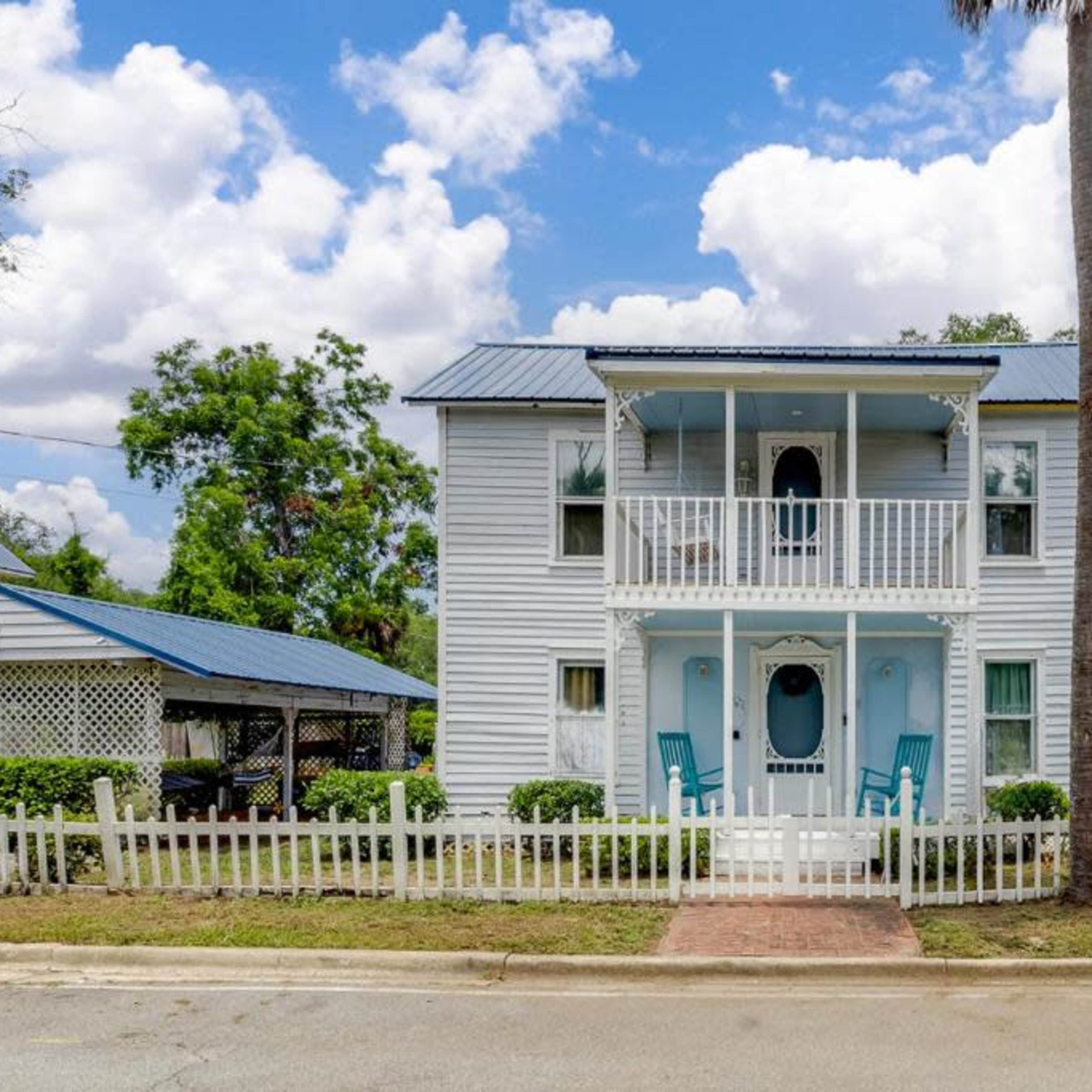 A two-story, light blue house with a white picket fence and a front porch featuring two chairs is situated along a road under a partly cloudy sky.
