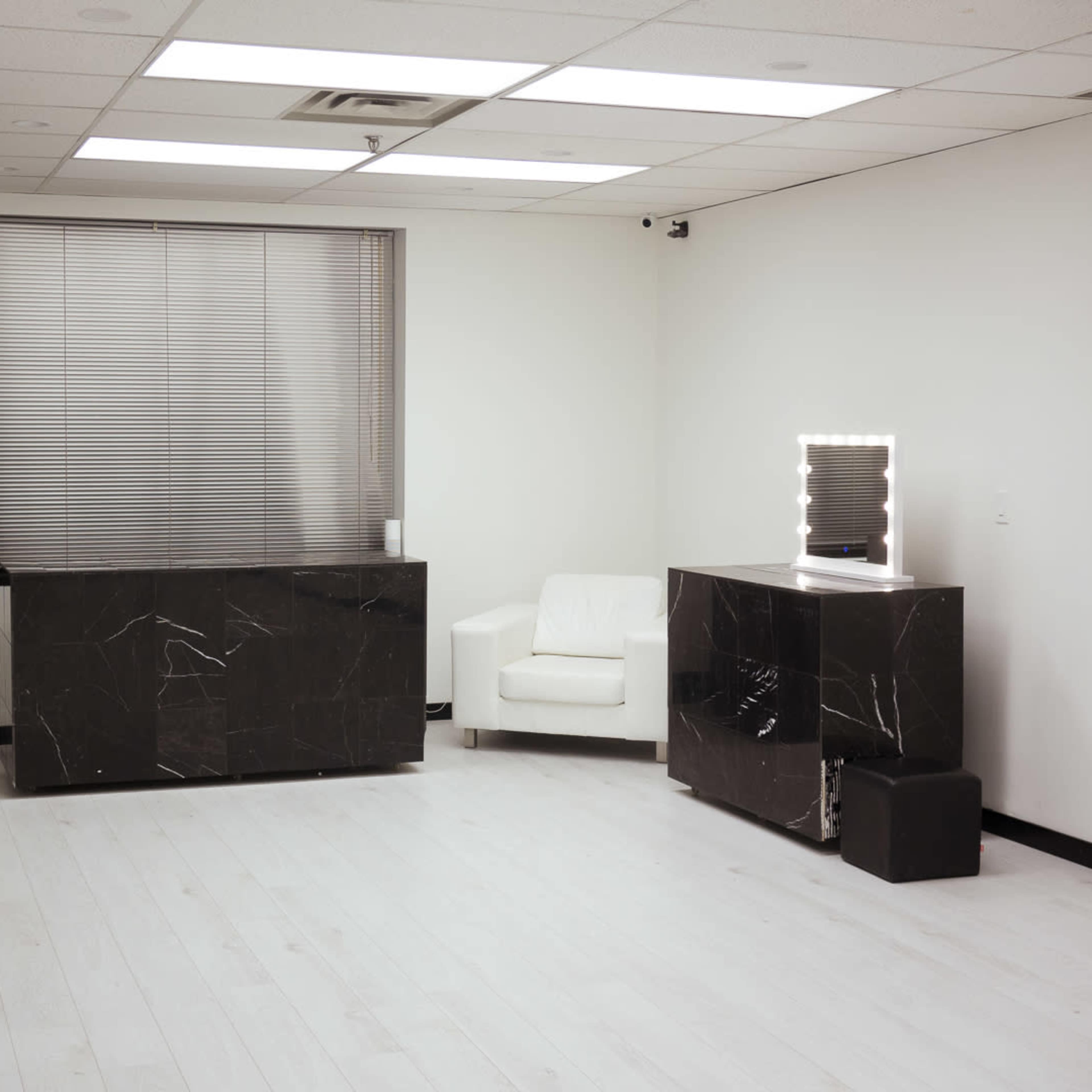 A minimalist room with a white floor, a large window with blinds, a potted plant, a black marble counter, a white armchair, and a backlit mirror on a second black marble counter.