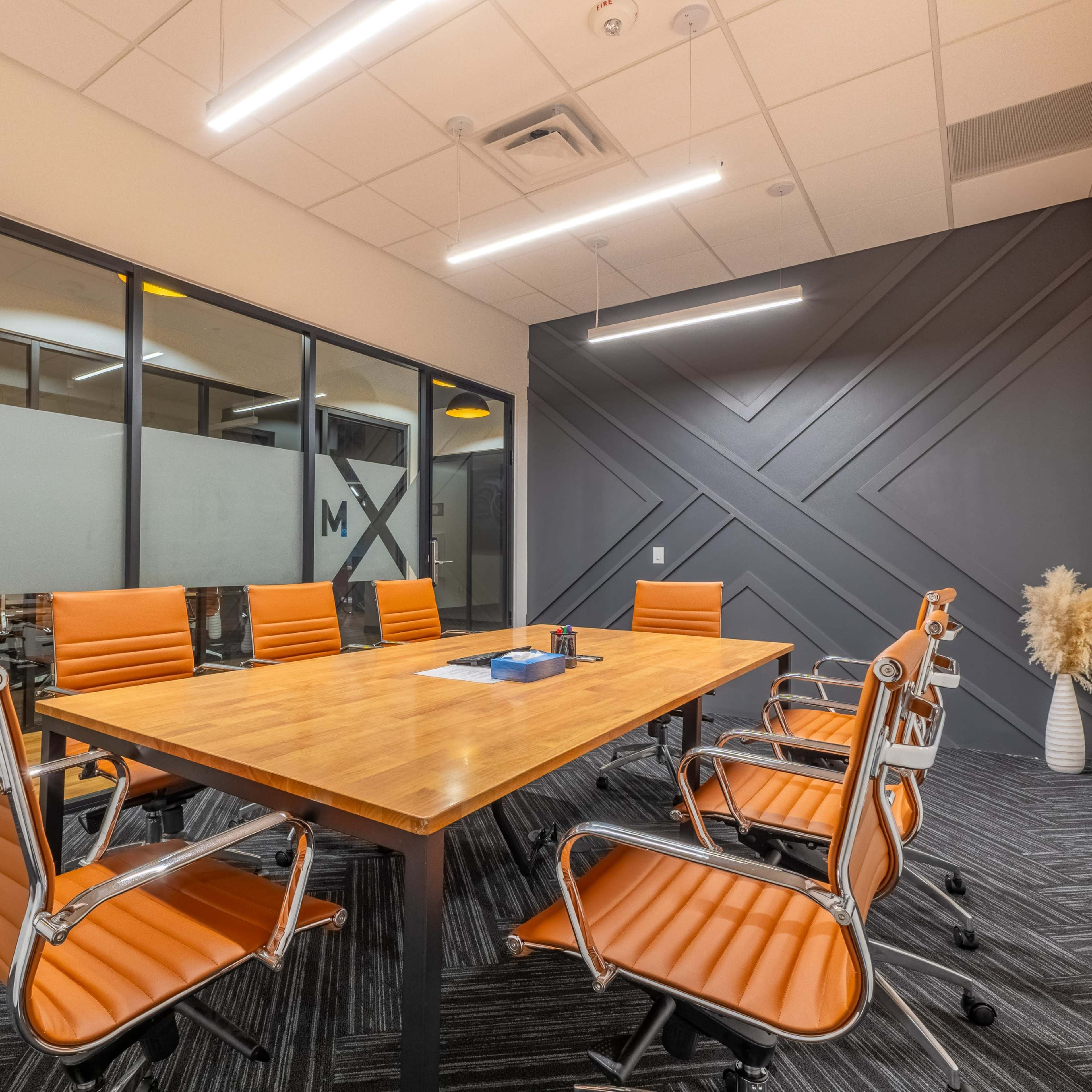 A modern conference room features a wooden table surrounded by orange leather chairs and large windows.