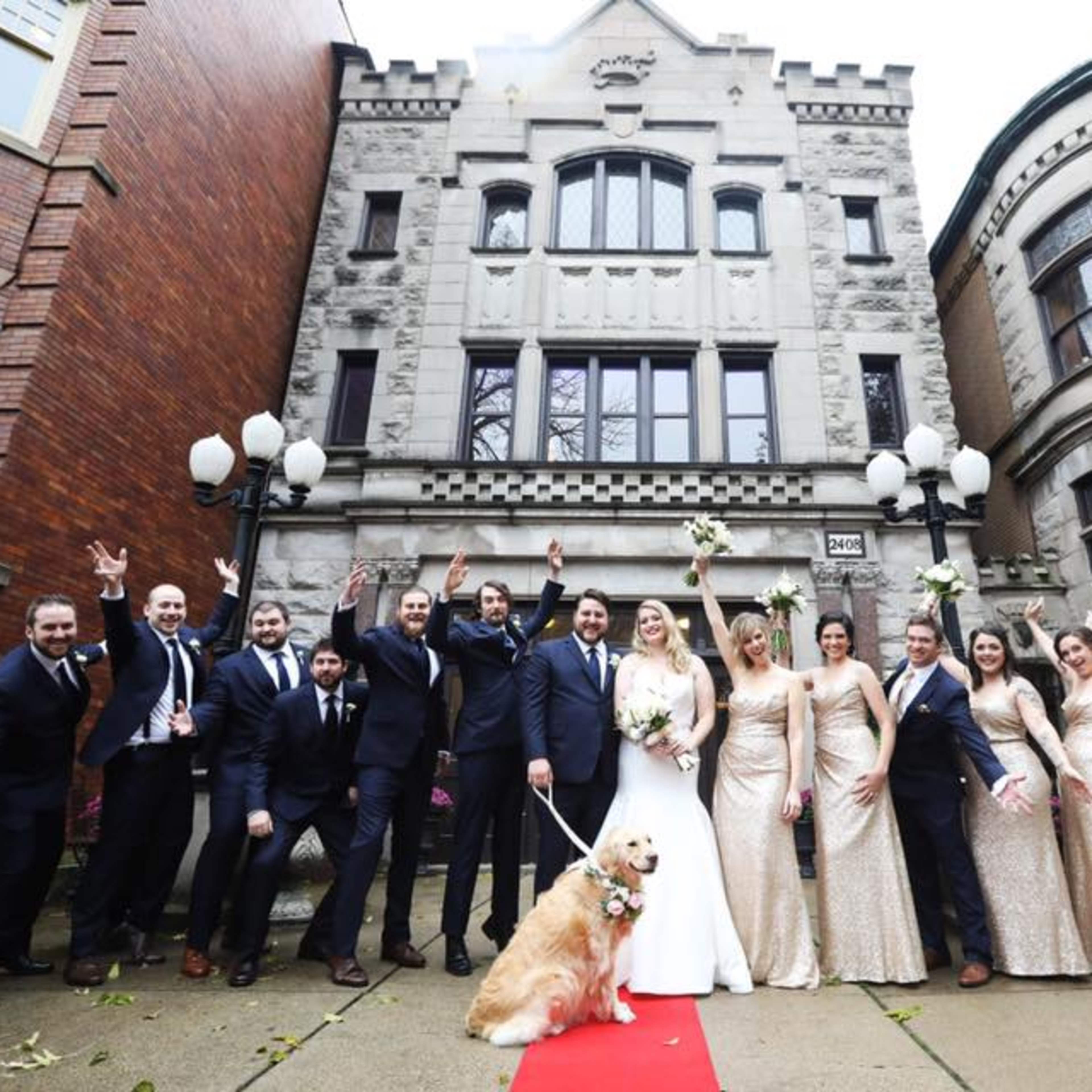 A wedding party poses joyfully outside a historic building, with the bride and groom in the center and a golden retriever at their feet, surrounded by bridesmaids in gold dresses and groomsmen in suits.