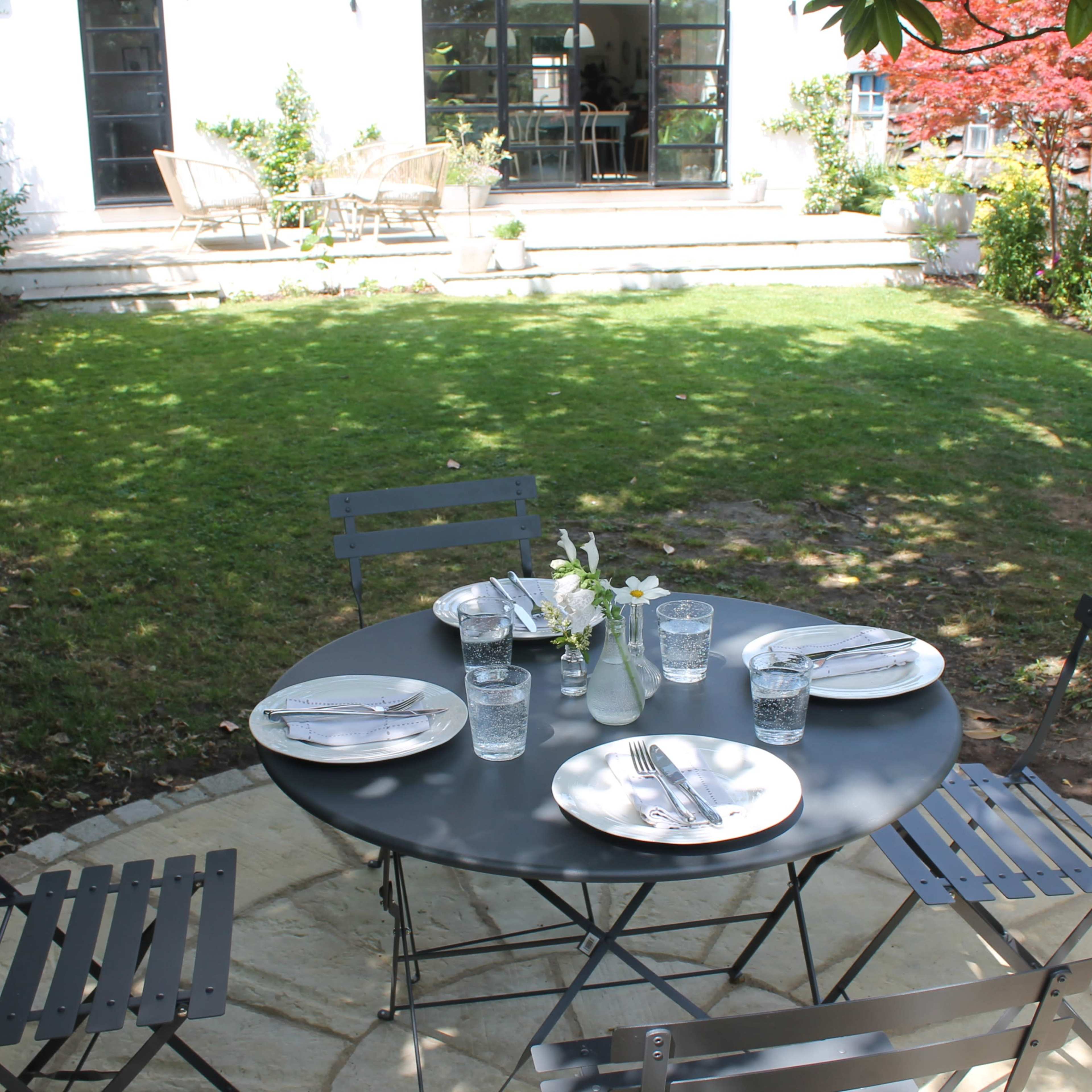 A round table with four chairs is set for dining in a grassy backyard, featuring a vase of flowers and plates at each place.