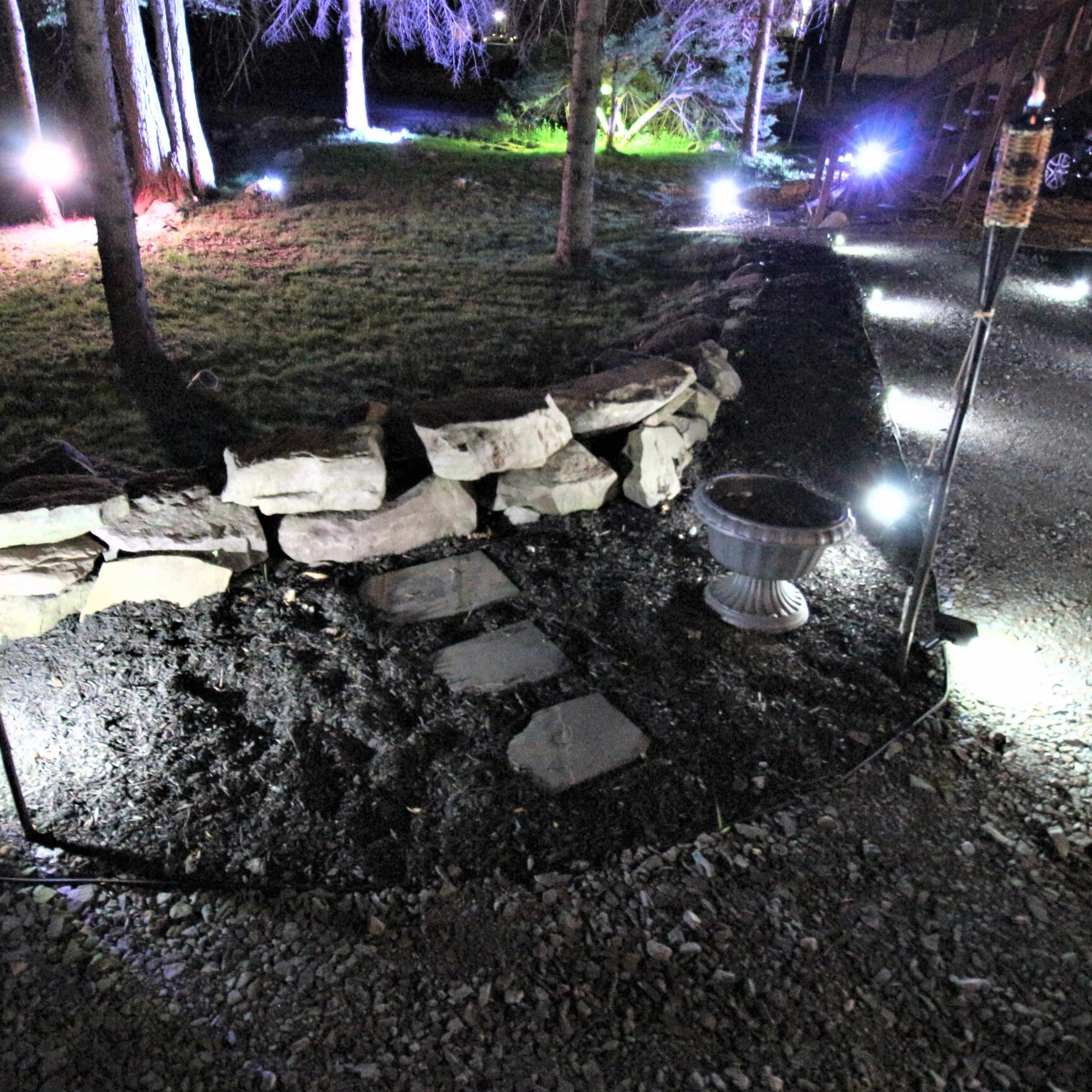 A landscaped outdoor area at night, featuring stone paths, ornamental lights, and torches along a gravel border.