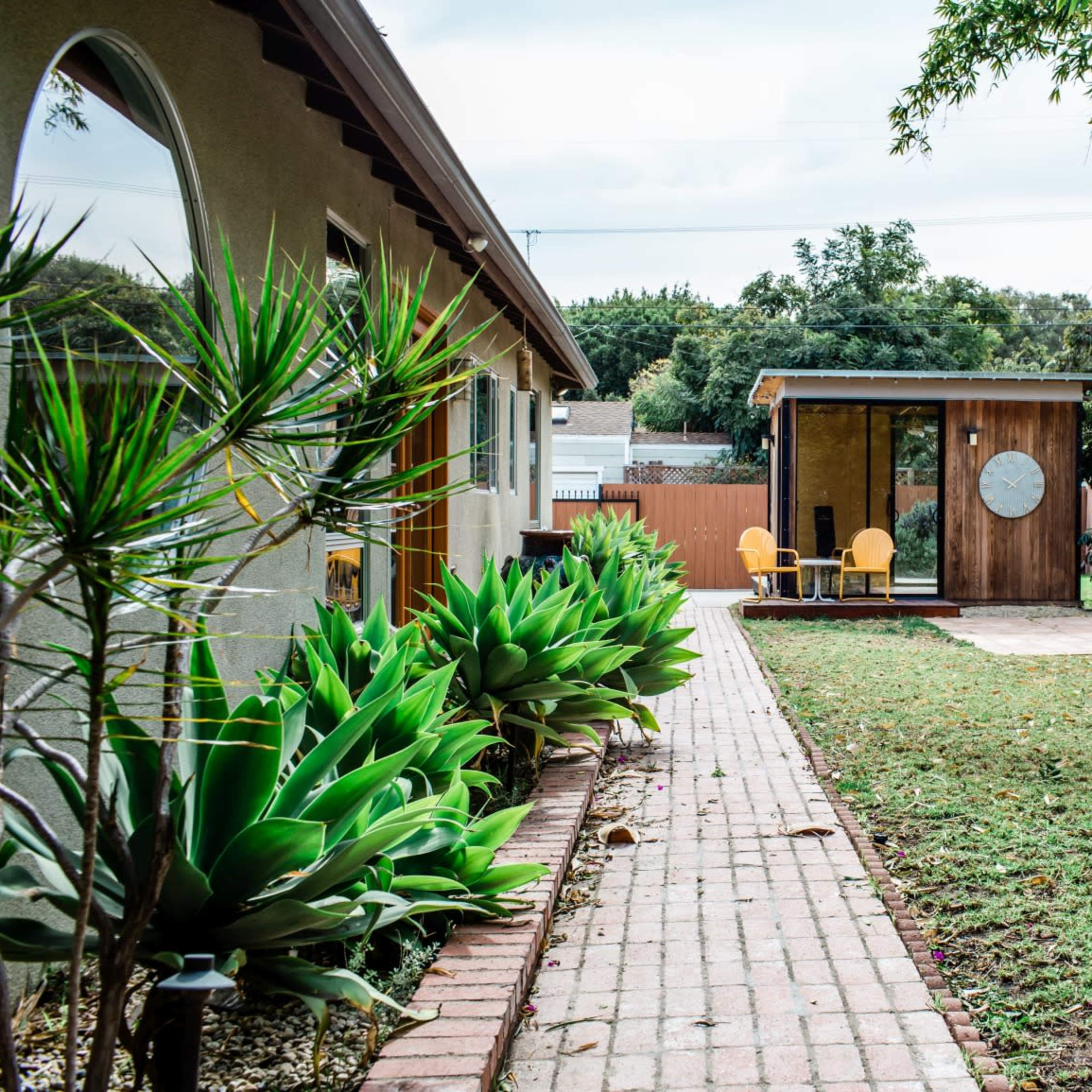 A series of brick pathways lead through a garden area with succulents, flanked by a modern shed and a small patio with chairs.