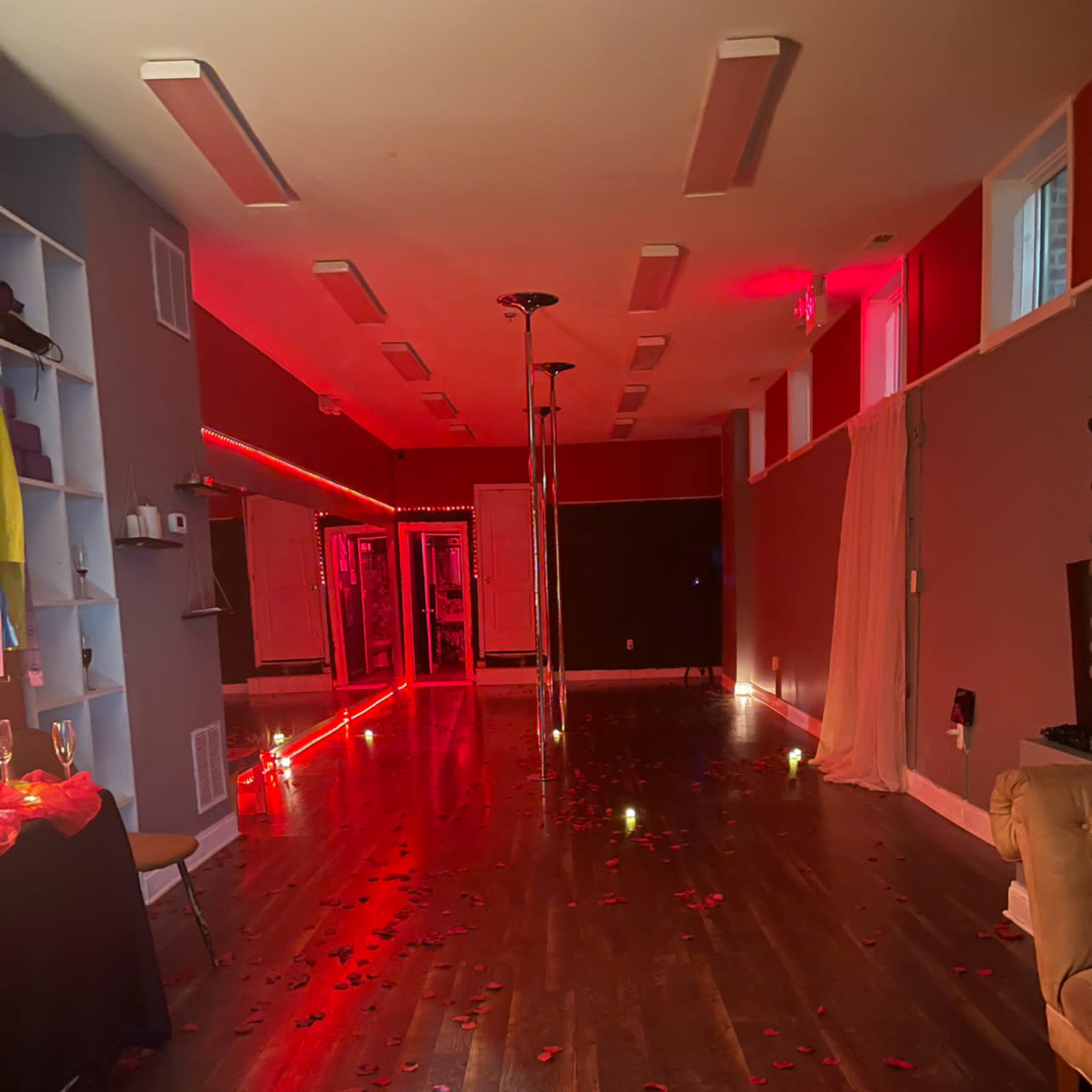 A dimly lit room with a dance pole at the center, adorned with rose petals on the wooden floor and illuminated by red lighting.