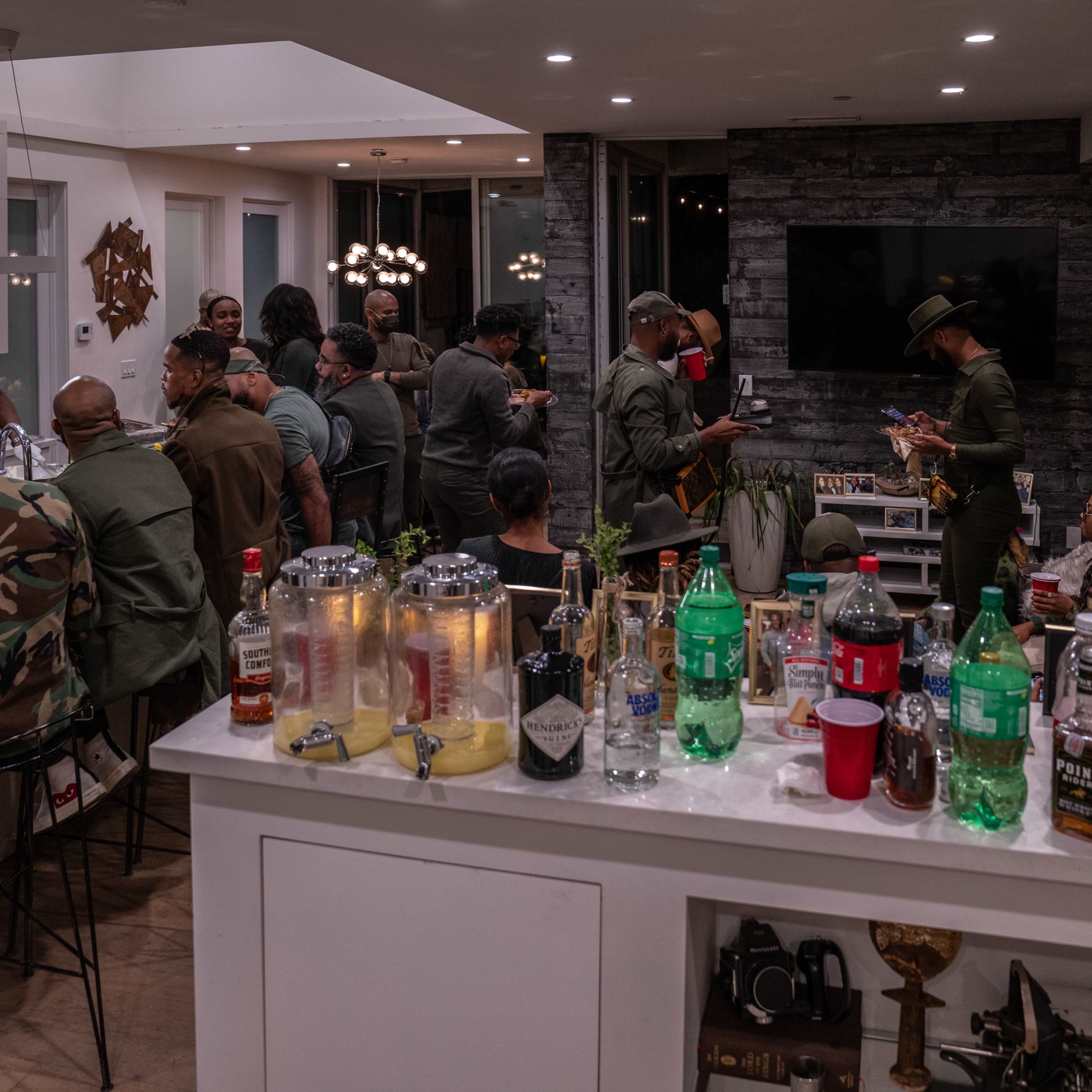 A diverse group of people gathers in a modern kitchen and dining area, engaged in conversation and food preparation, with various drinks displayed on the counter.