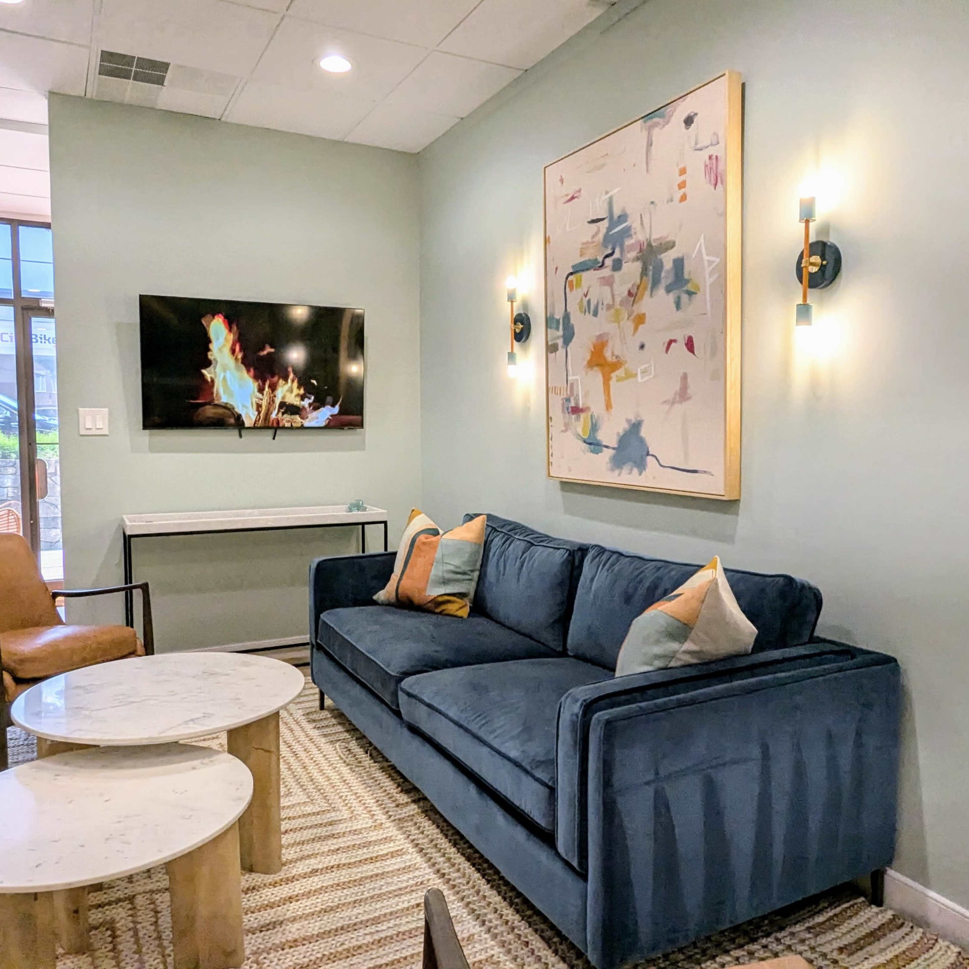 A modern sitting area with a blue couch, two coffee tables, a wall-mounted television, and a piece of abstract art on the wall.
