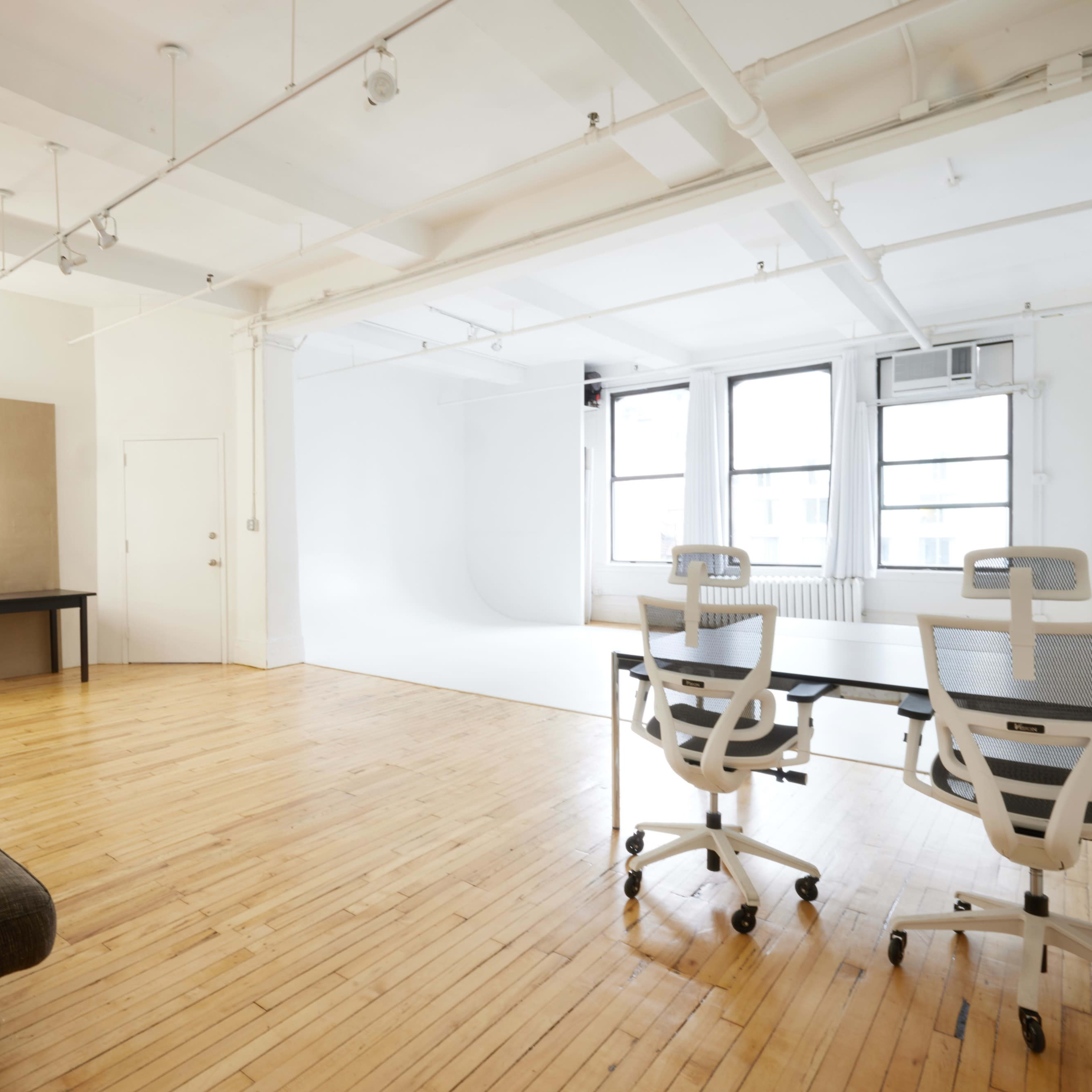 A spacious, well-lit studio featuring a seating area with a couch, a dressing table with lights, and two office chairs positioned at a table.